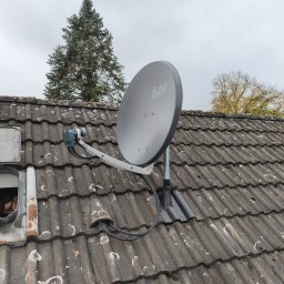 Im-Sat Dortmund - Szara antena satelitarna marki Fuba zamontowana na brudnym, szarym dachu z dachówki ceramicznej. W tle widoczne drzewa i pochmurne niebo.