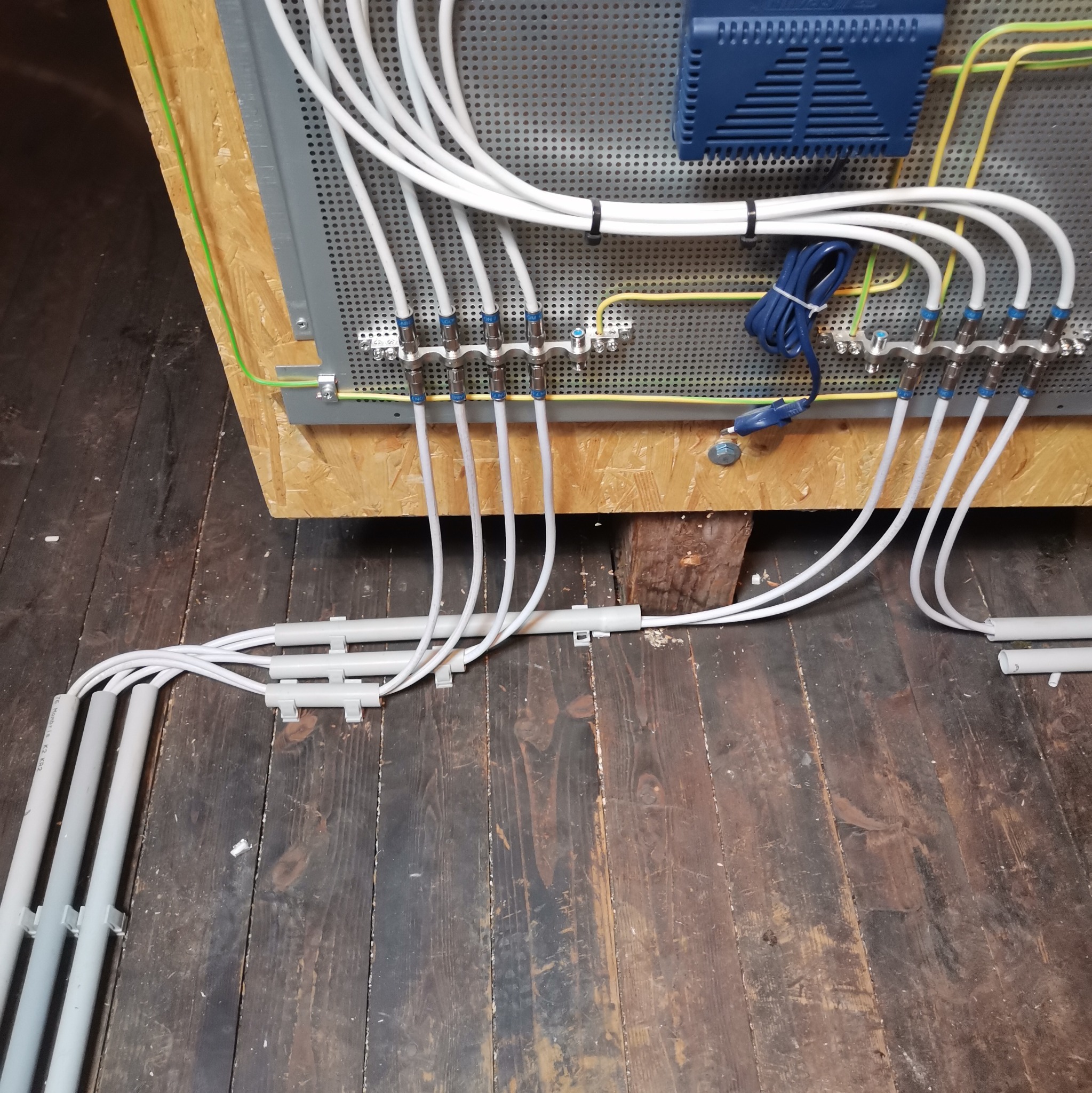 Rozdzielacz sygnału z licznymi kablami koncentrycznymi, zamontowany na płycie OSB z uziemieniem, widoczne kable w peszlach na podłodze z desek.