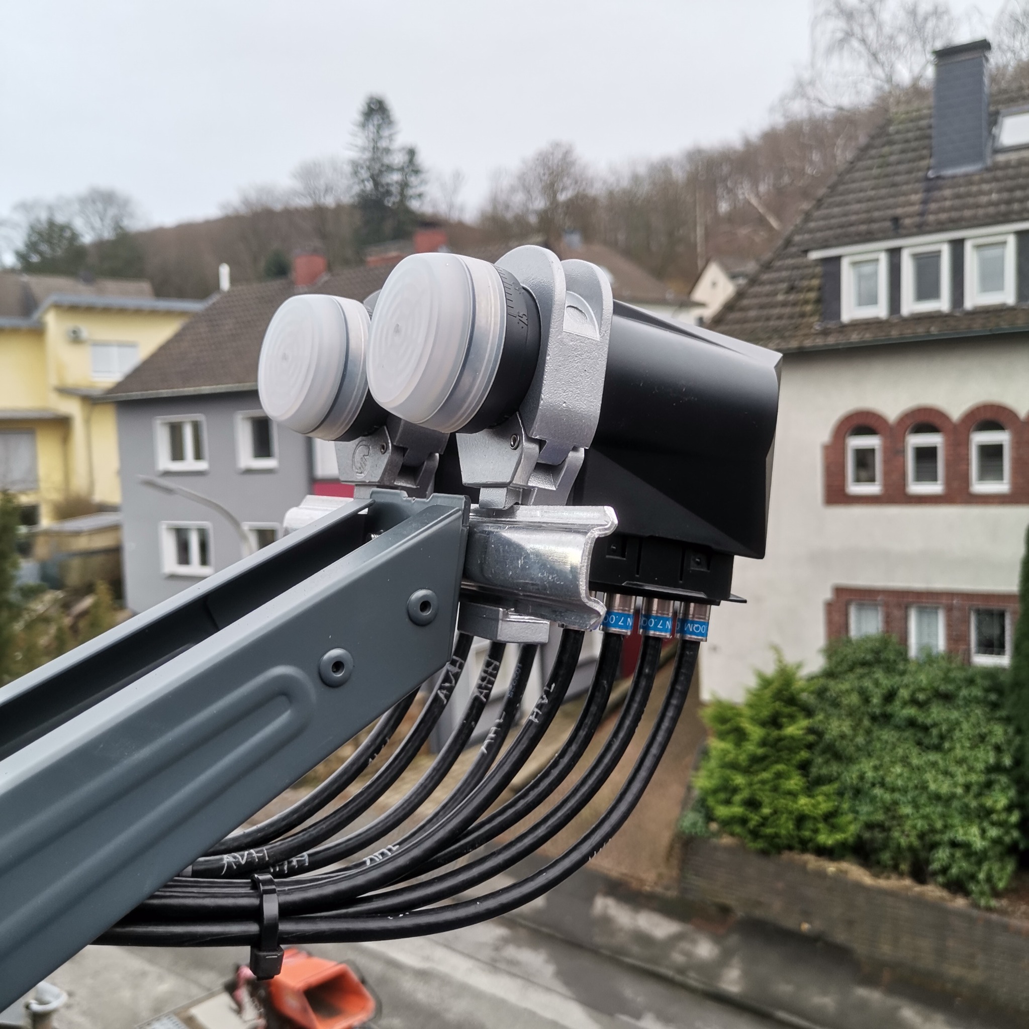 Zewnętrzna instalacja satelitarna z dwoma konwerterami LNB, widoczne kable antenowe z oznaczeniami, na tle zabudowa jednorodzinna w pochmurny dzień.