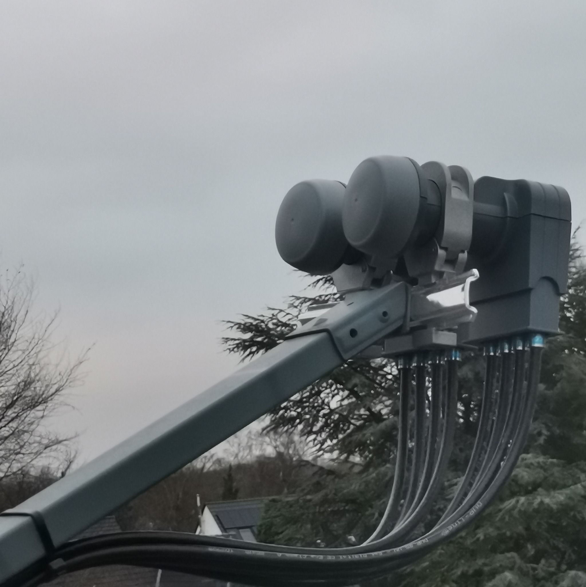 Szary uchwyt antenowy z czterema konwerterami i licznymi przewodami, na tle pochmurnego nieba i drzew.