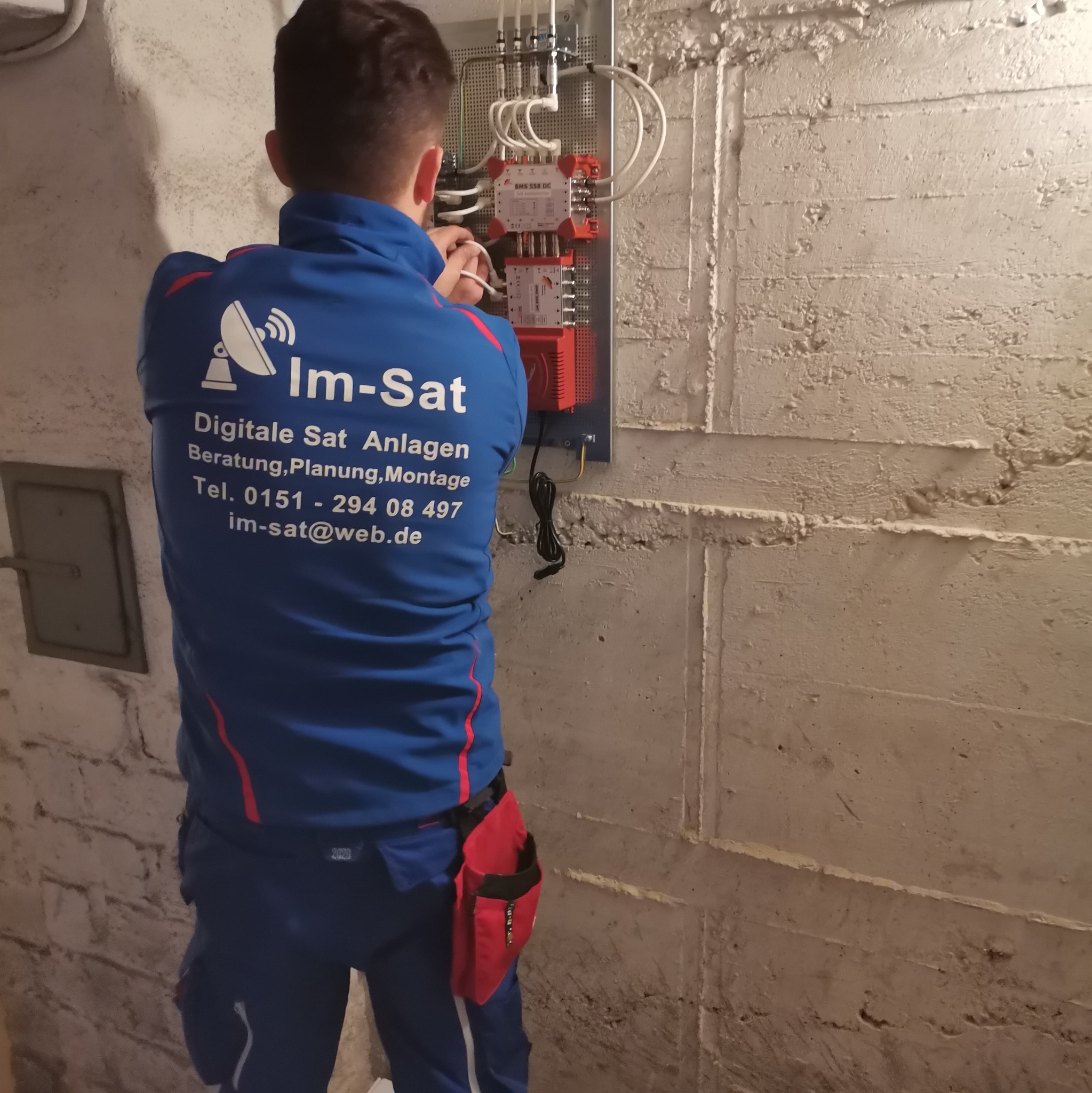 Instalator w niebieskim uniformie firmy Im-Sat podłącza kable do rozdzielnicy sygnału satelitarnego na szorstkiej, pomalowanej ścianie w piwnicy.