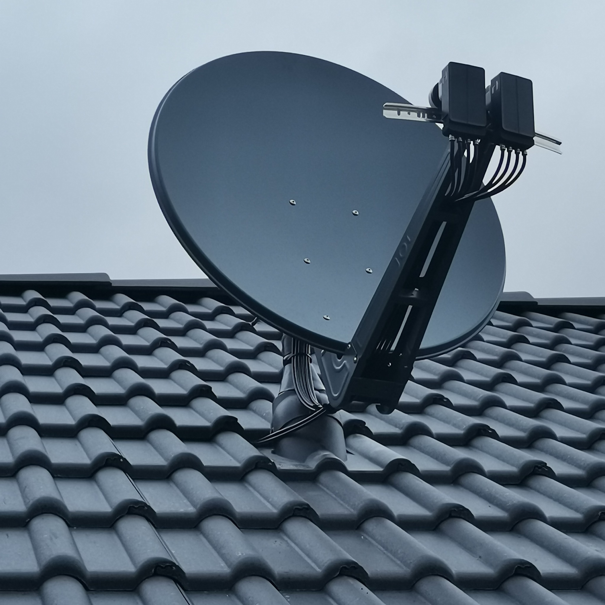 Czarna antena satelitarna zamontowana na szarym dachu pokrytym ceramiczną dachówką, widoczne kable i elementy mocujące.