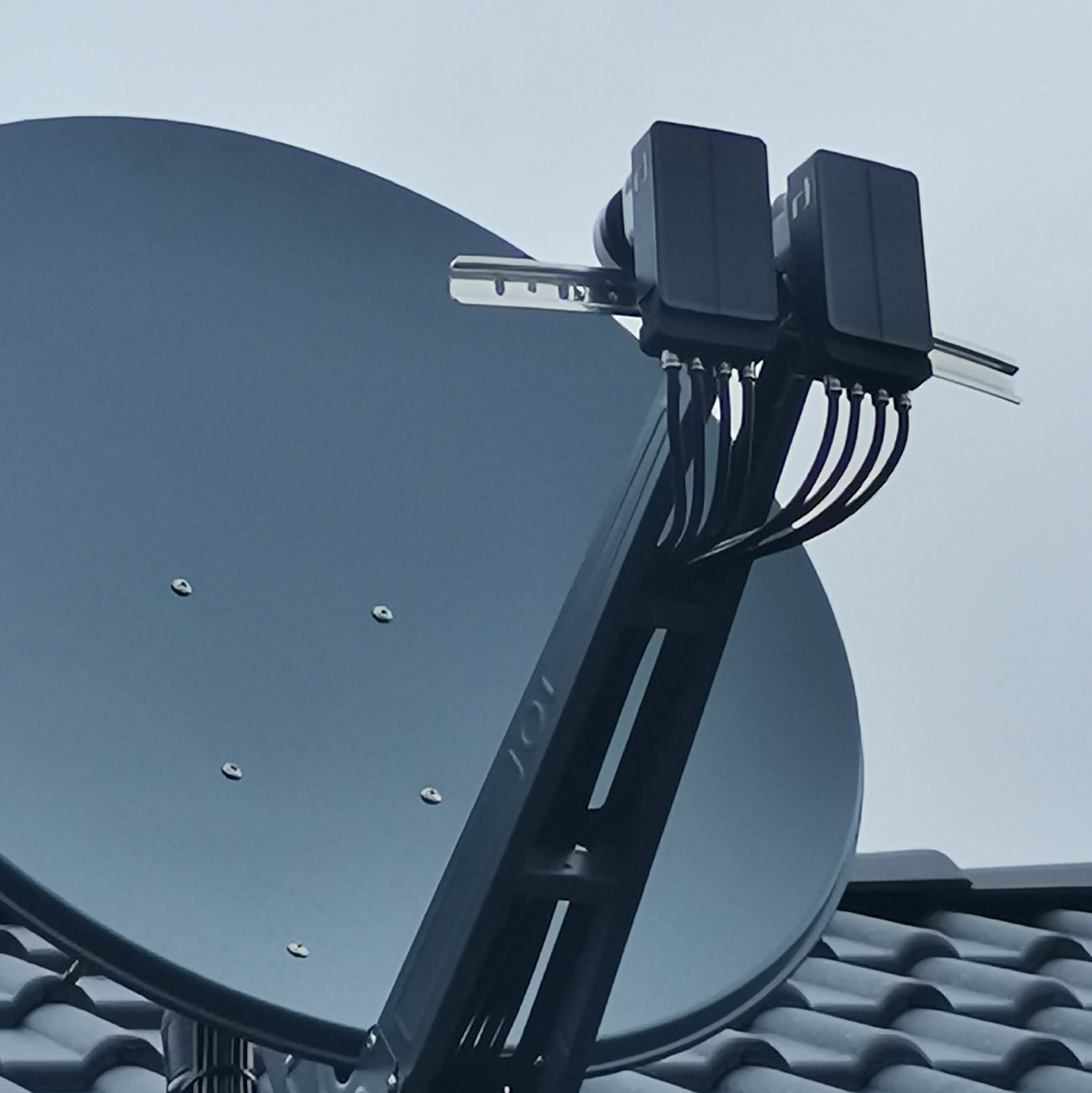 Czarna antena satelitarna z dwoma konwerterami na dachu pokrytym szarą dachówką, widok z dołu na pochmurne niebo.