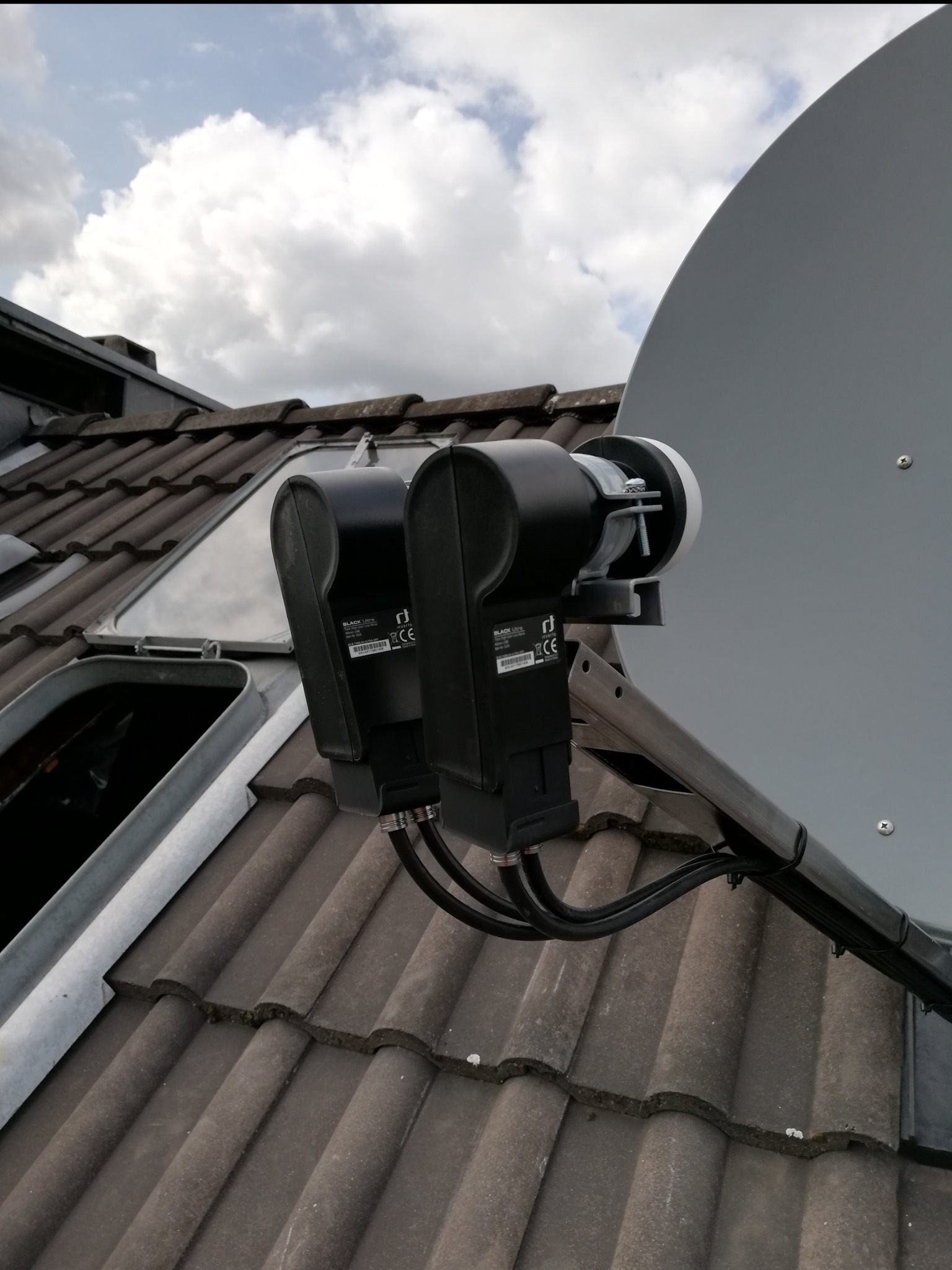 Antena satelitarna z dwoma konwerterami typu monoblock zamontowana na dachu pokrytym brązową dachówką, widok na pochmurne niebo.