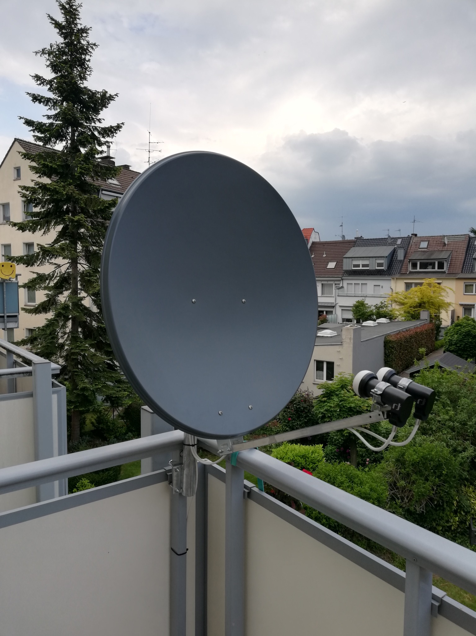 Szara antena satelitarna zamontowana na balkonie bloku mieszkalnego, widok na okoliczne budynki i zieleń, pochmurne niebo w tle.