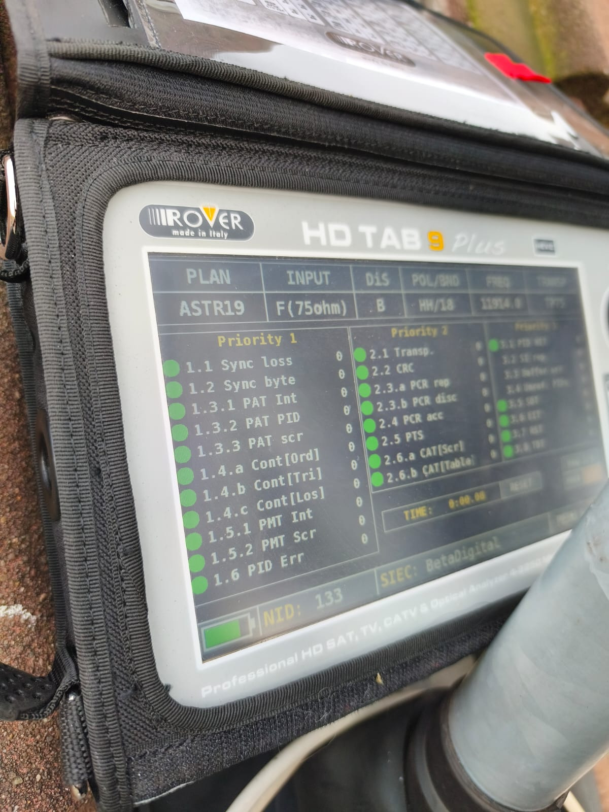 Miernik sygnału satelitarnego Rover HD TAB 9 Plus w czarnym futerale, wyświetlający parametry sygnału ASTR19. Widoczne zielone wskaźniki priorytetów i wartości parametrów.