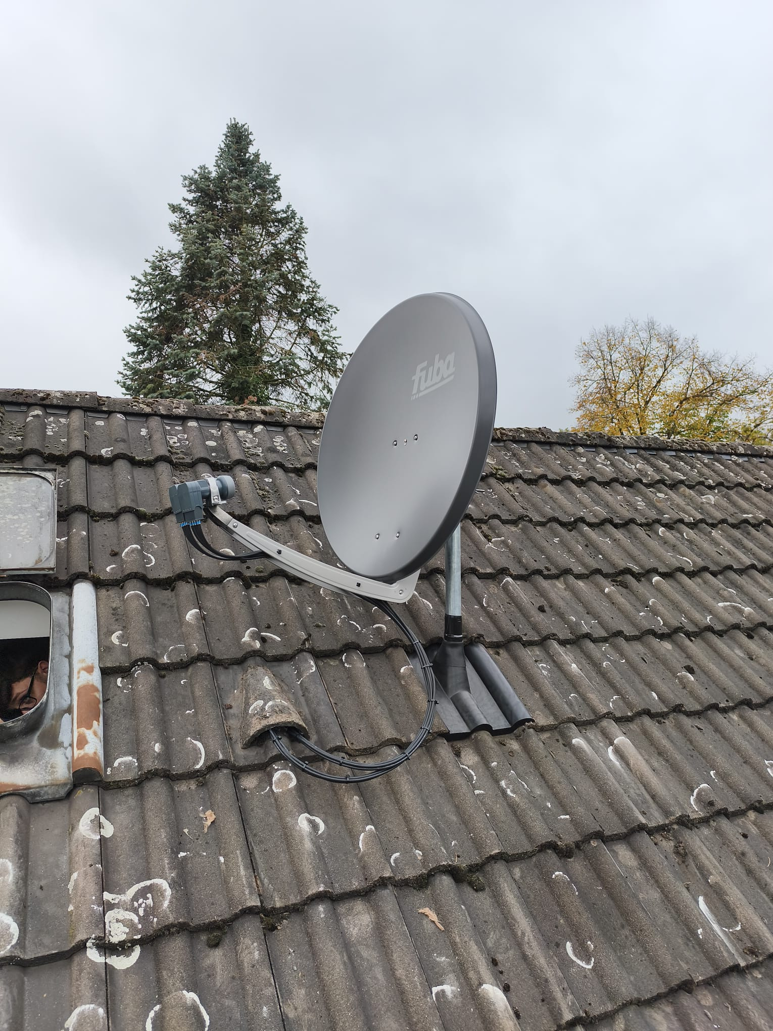 Szara antena satelitarna marki Fuba zamontowana na brudnym, szarym dachu z dachówki ceramicznej. W tle widoczne drzewa i pochmurne niebo.