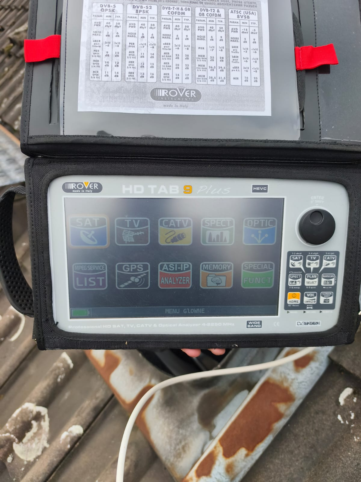 Miernik sygnału satelitarnego Rover HD Tab 9 Plus w czarnym futerale, leżący na dachu. Ekran urządzenia wyświetla menu z ikonami SAT, TV, CATV, SPECT i OPTIC.