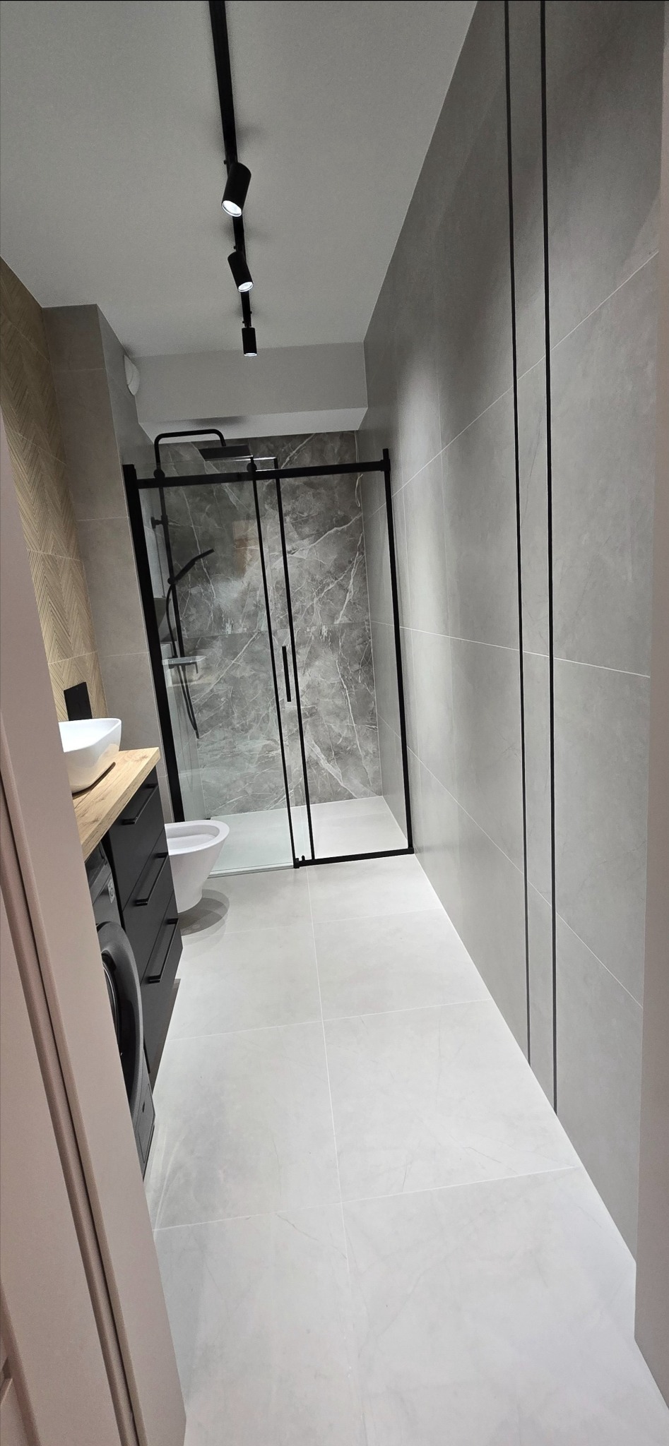 Nowoczesna łazienka z szarymi płytkami, kabiną prysznicową z czarną ramą, umywalką nablatową i oświetleniem punktowym.