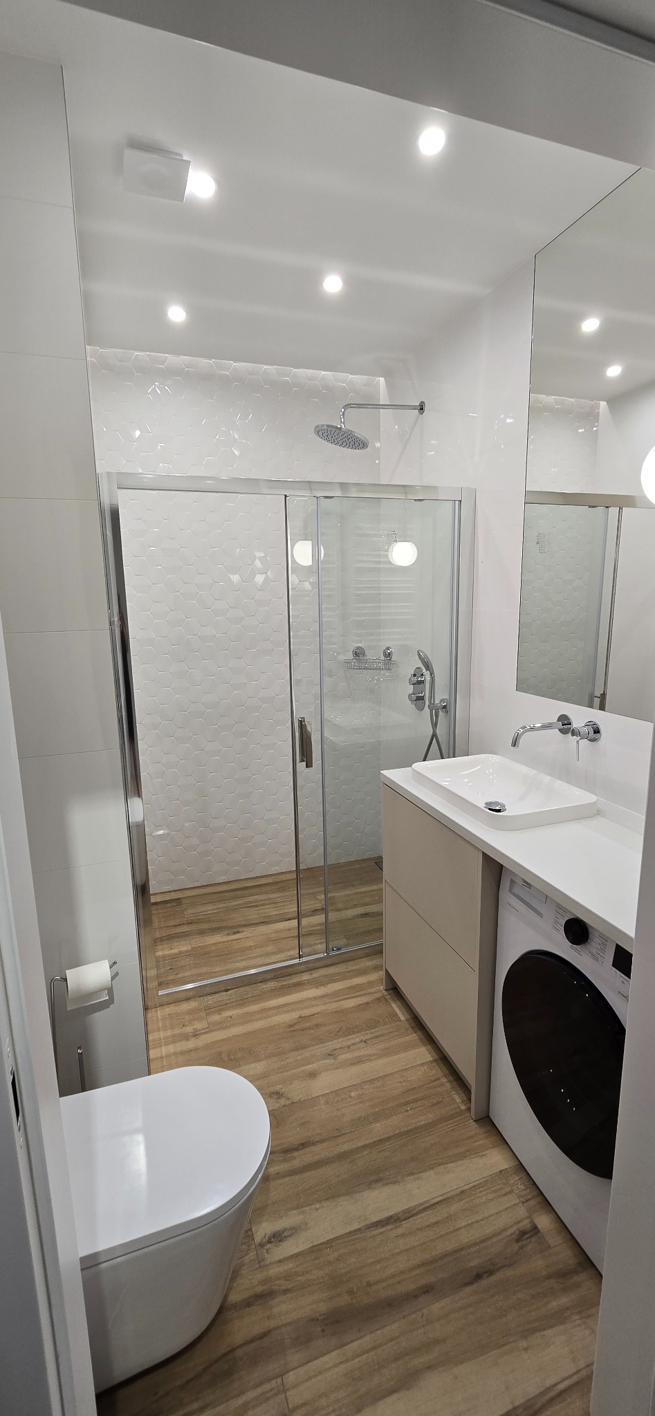 Nowoczesna łazienka z kabiną prysznicową, umywalką nablatową na szafce z pralką w zabudowie, toaletą i podłogą imitującą drewno.