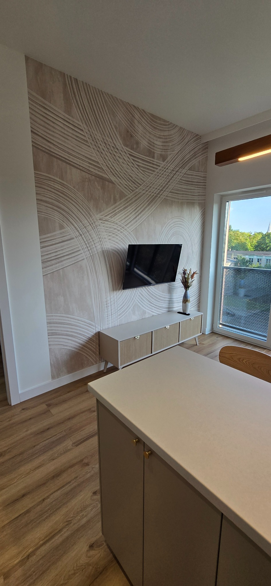 Salon po metamorfozie z dekoracyjną tapetą w beżowe fale, wiszącym telewizorem, szafką RTV z jasnego drewna i minimalistycznym wazonem z suszonymi kwiatami.