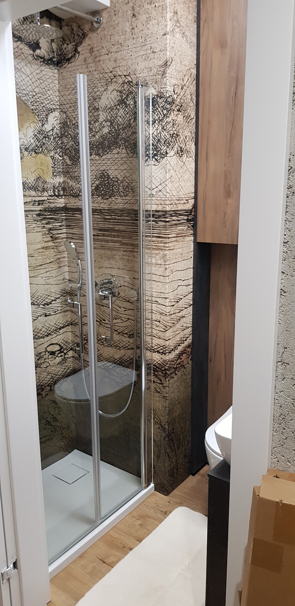 Kabina prysznicowa z grafiką na ścianie, chromowana armatura, biały brodzik i drewnopodobna szafka w nowocześnie urządzonej łazience.