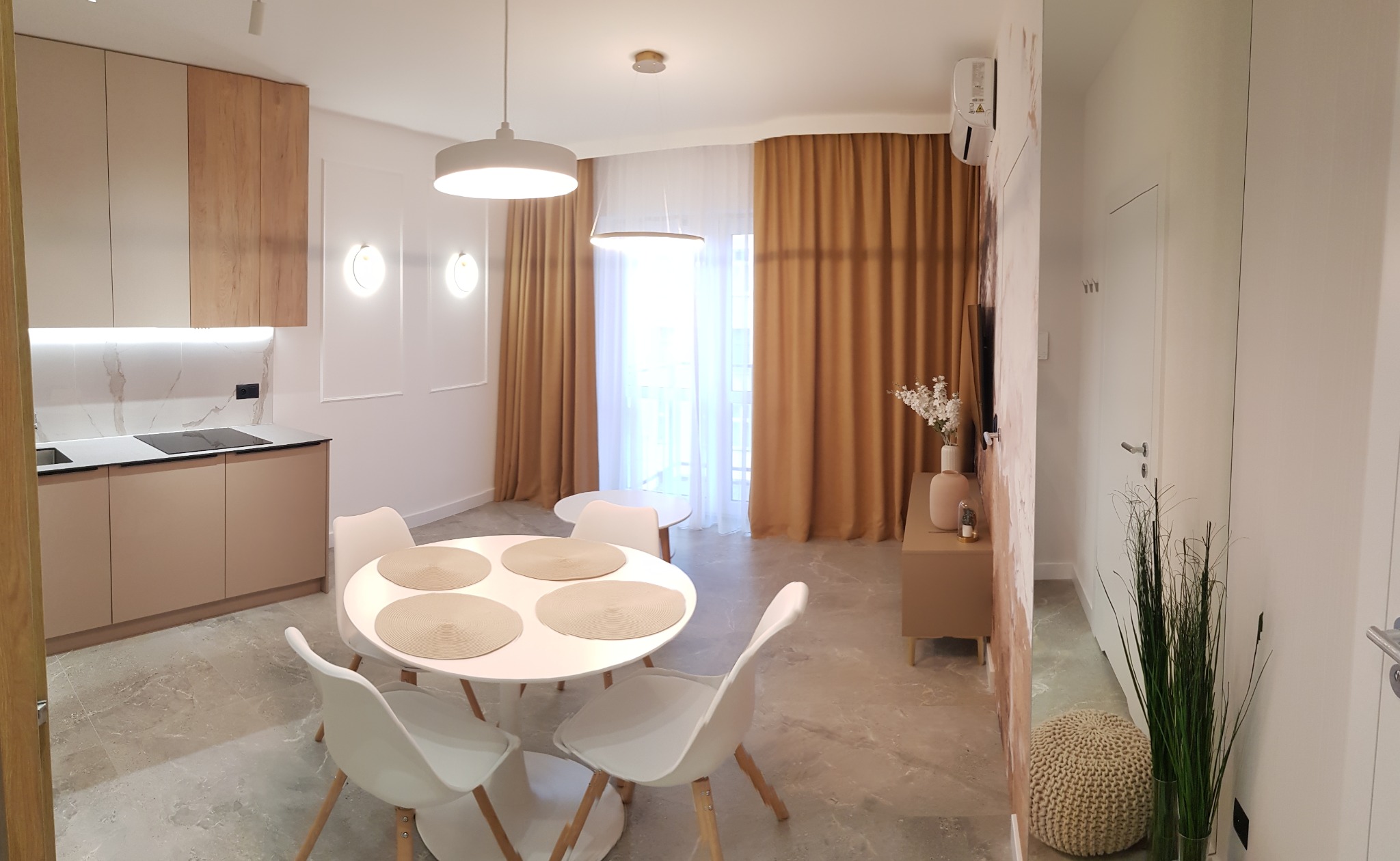 Jasne, nowocześnie urządzone wnętrze mieszkania z aneksem kuchennym, okrągłym stołem z czterema krzesłami, złotymi zasłonami i dekoracyjną trawą w wazonie przy ścianie.