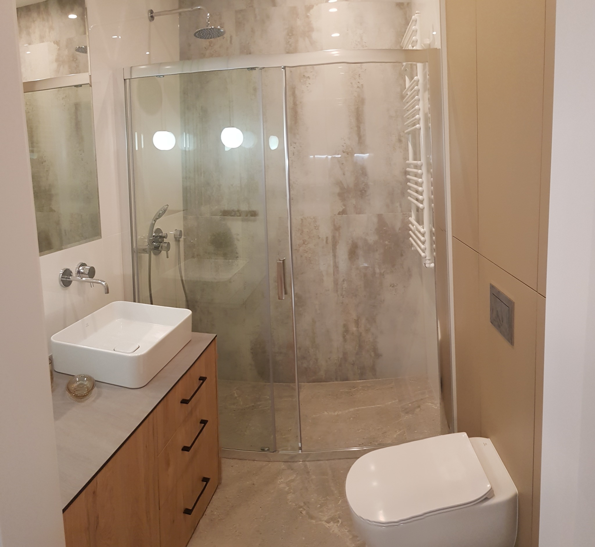 Nowoczesna łazienka po remoncie z kabiną prysznicową ze szklanymi drzwiami przesuwnymi, umywalką nablatową na szafce z drewnopodobnym wykończeniem i wiszącą toaletą.