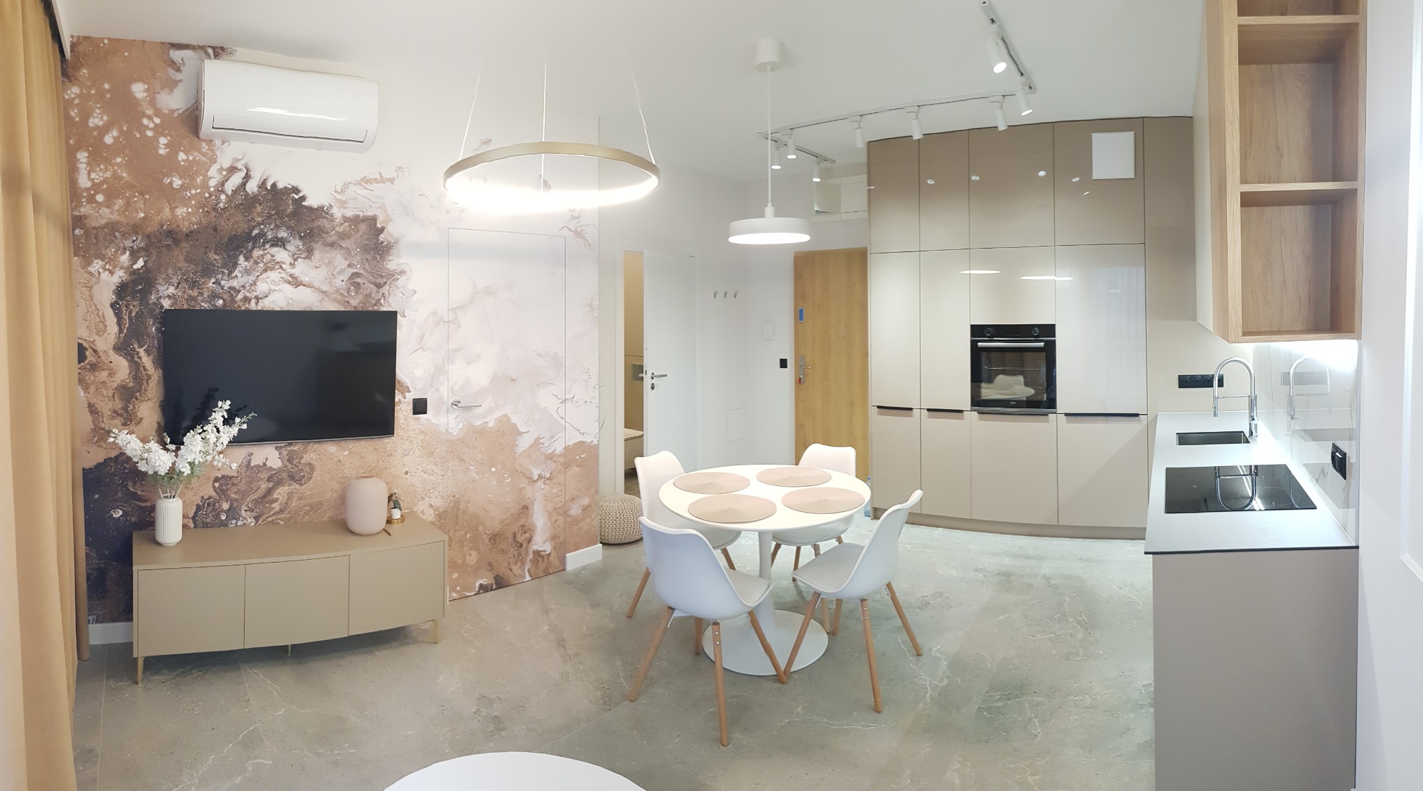 Nowoczesny salon z aneksem kuchennym w odcieniach beżu i bieli, ściana z abstrakcyjnym wzorem przypominającym marmur, okrągły stół z białymi krzesłami, minimalistyczne oświetlenie.