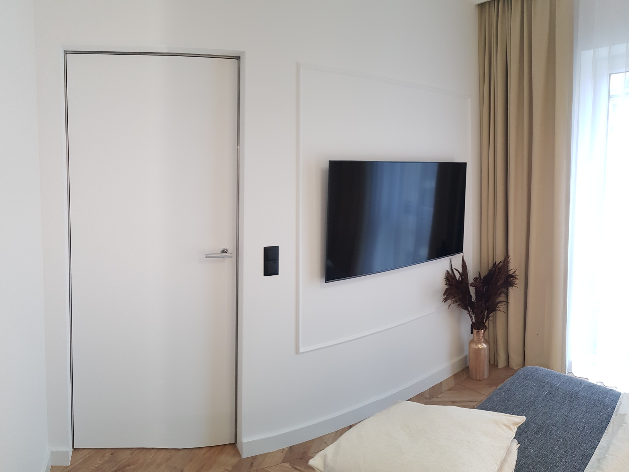 Minimalistyczne wnętrze: białe drzwi bezprzylgowe z satynową klamką, telewizor na ścianie z dekoracyjną ramą, złoty wazon z suszem, drewniana podłoga i beżowe zasłony.