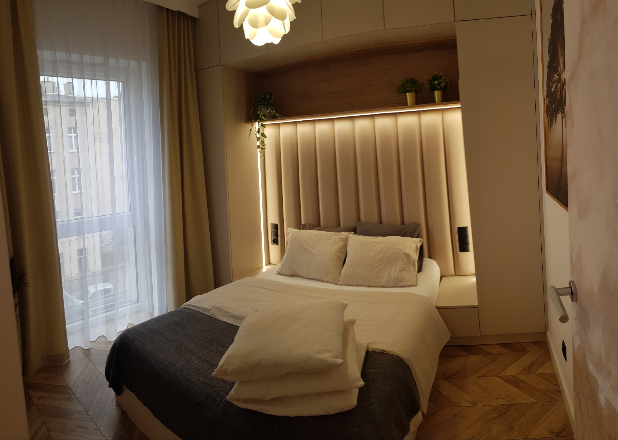 Nowoczesna sypialnia z beżowym tapicerowanym zagłówkiem, drewnianą półką z roślinami, designerską lampą i parkietem w jodełkę