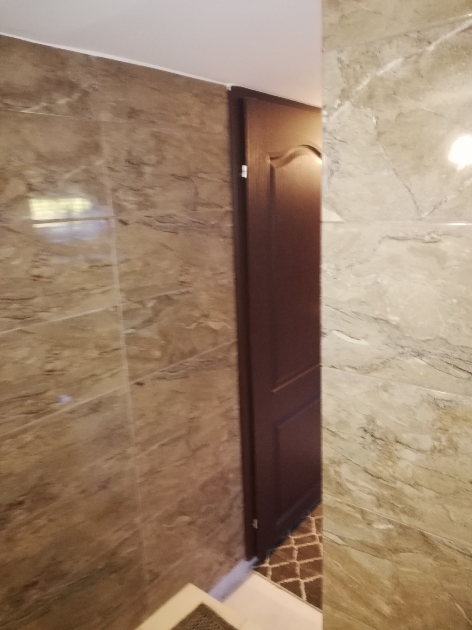 Fragment łazienki po remoncie: ściany wyłożone brązowo-beżowymi płytkami imitującymi marmur, widoczny fragment brązowych drzwi i wzorzysta podłoga w tle.