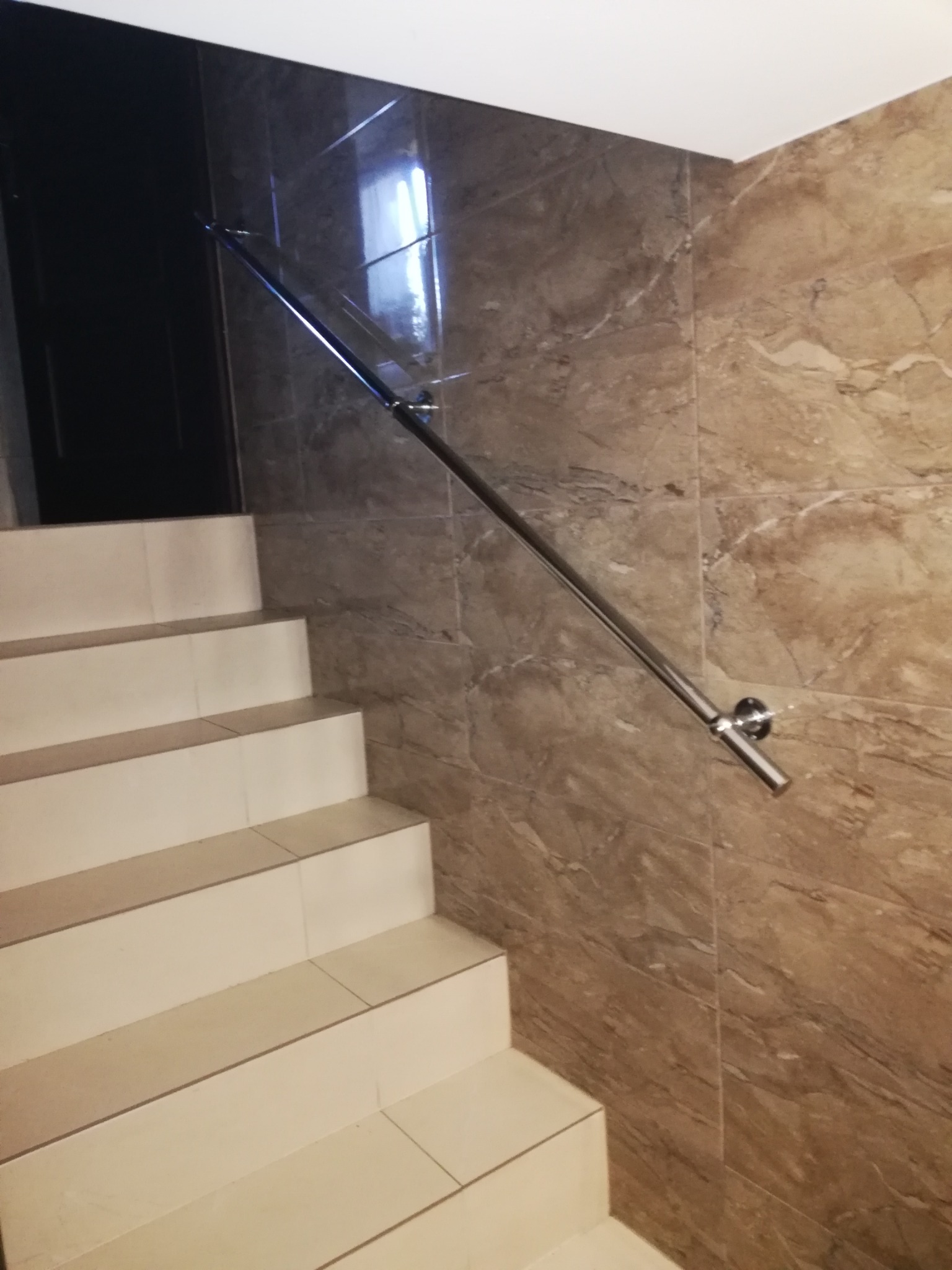 Schody wyłożone jasnymi płytkami ceramicznymi z metalową poręczą zamontowaną na ścianie z płytek imitujących marmur w odcieniach brązu i beżu, widoczny fragment czarnych drzwi.