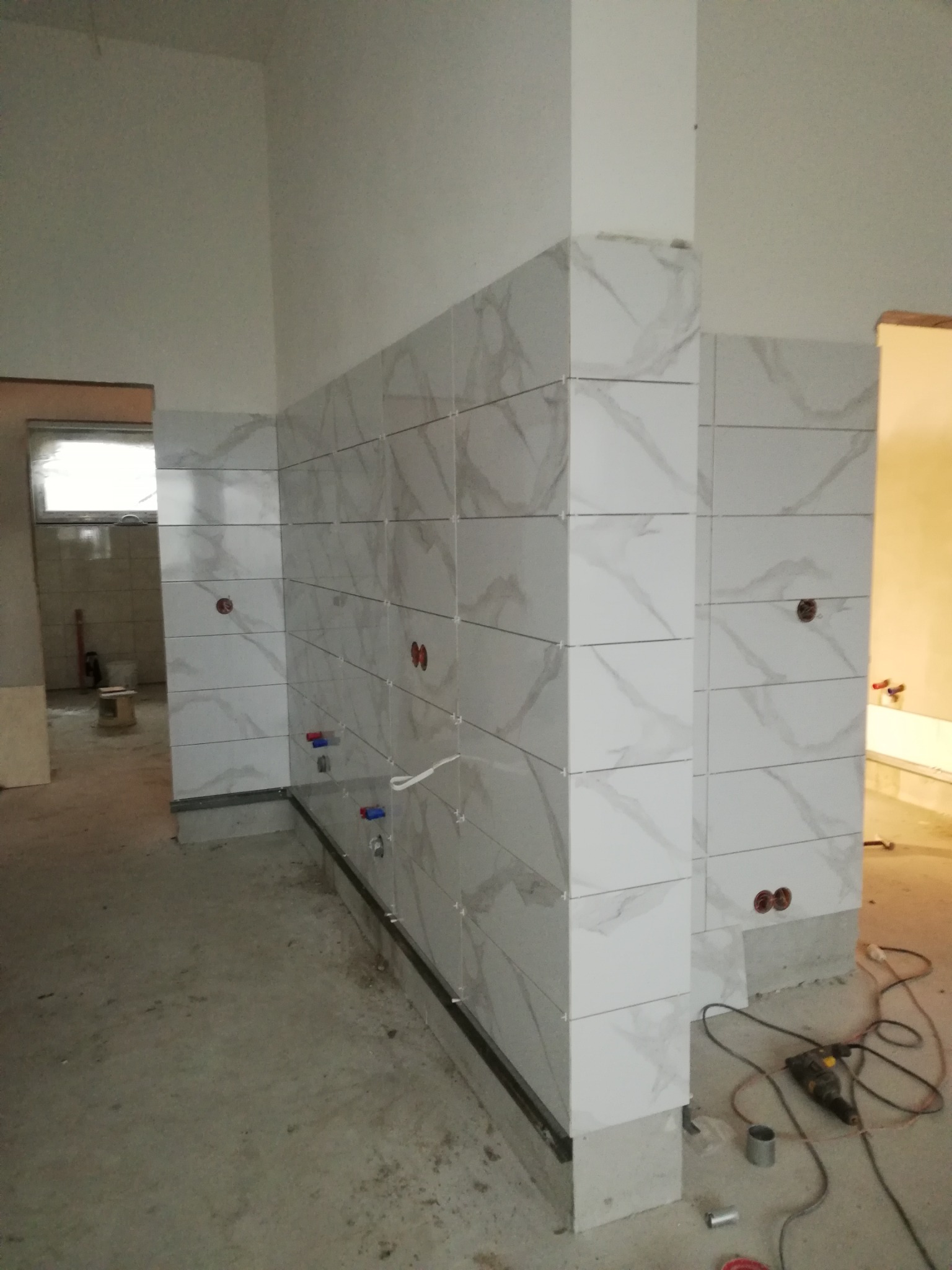 Ściana w trakcie układania białych płytek imitujących marmur, widoczne otwory na instalacje, narzędzia pracy i surowy beton na podłodze.