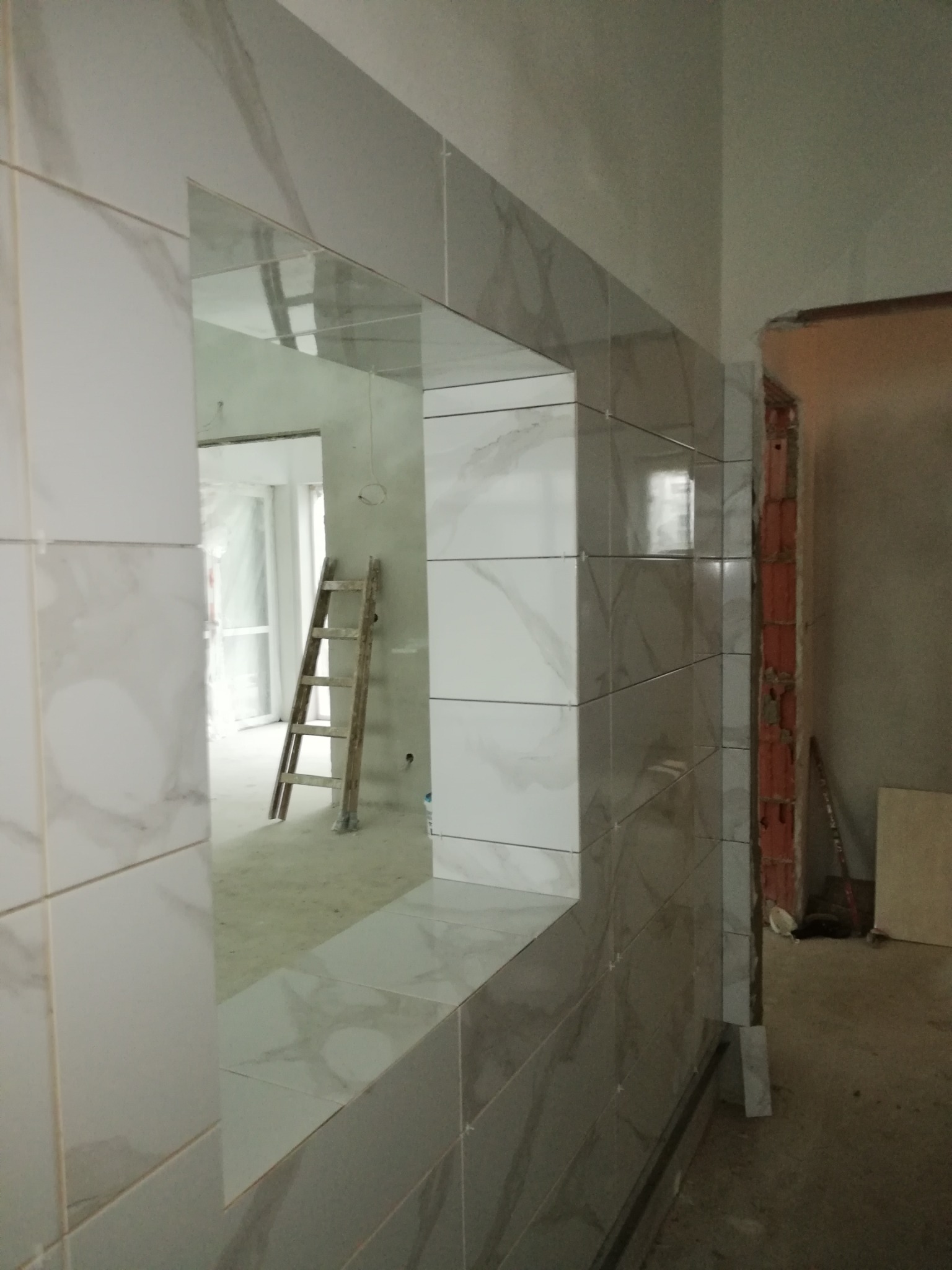 Ściana z ułożonymi białymi płytkami imitującymi marmur, z widocznym otworem przejściowym, w tle drabina oparta o ścianę, etap prac wykończeniowych.