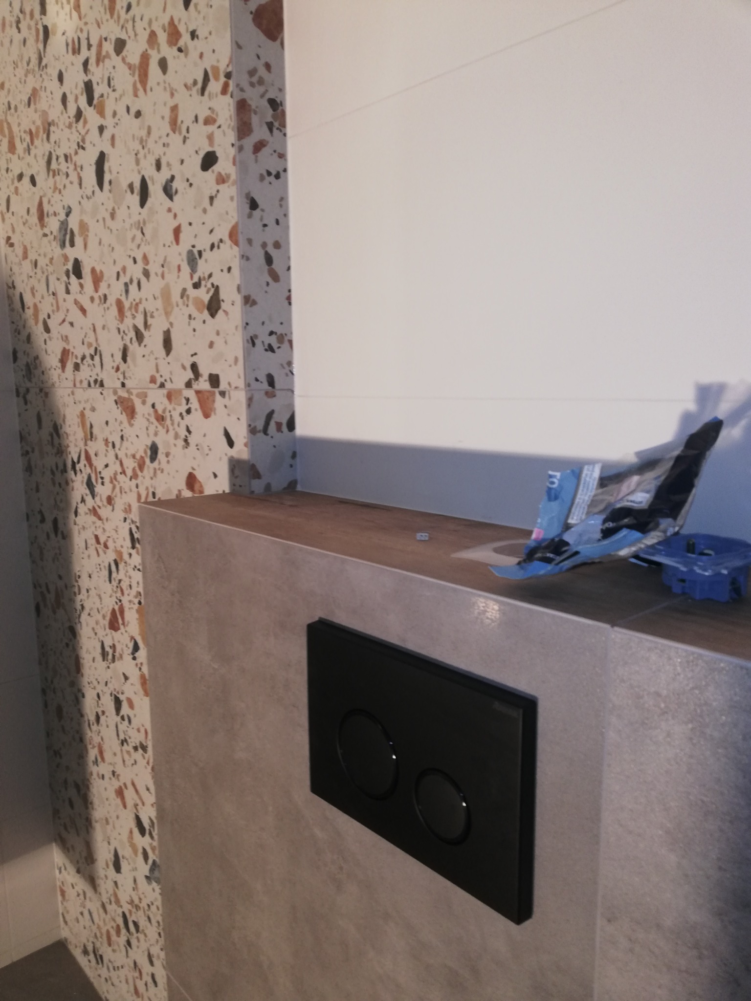 Ściana łazienki z płytkami imitującymi lastryko, zabudowa stelaża WC z czarnym panelem przycisków, resztki opakowań i narzędzi na blacie.