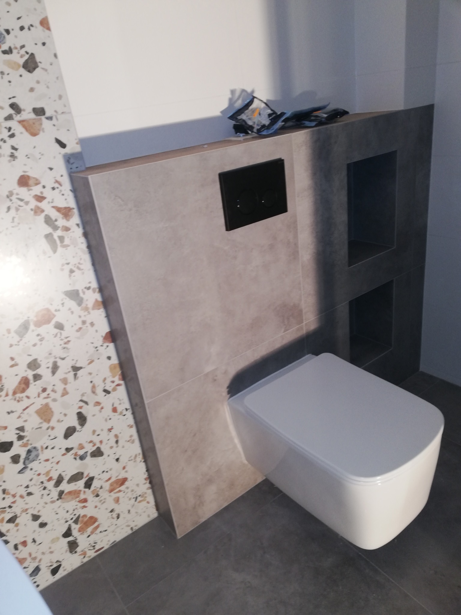 Podwieszana toaleta z czarnym przyciskiem spłukującym, zabudowana szarą płytką z wnękami, obok ściana z mozaiką lastryko, na górze leżą narzędzia.