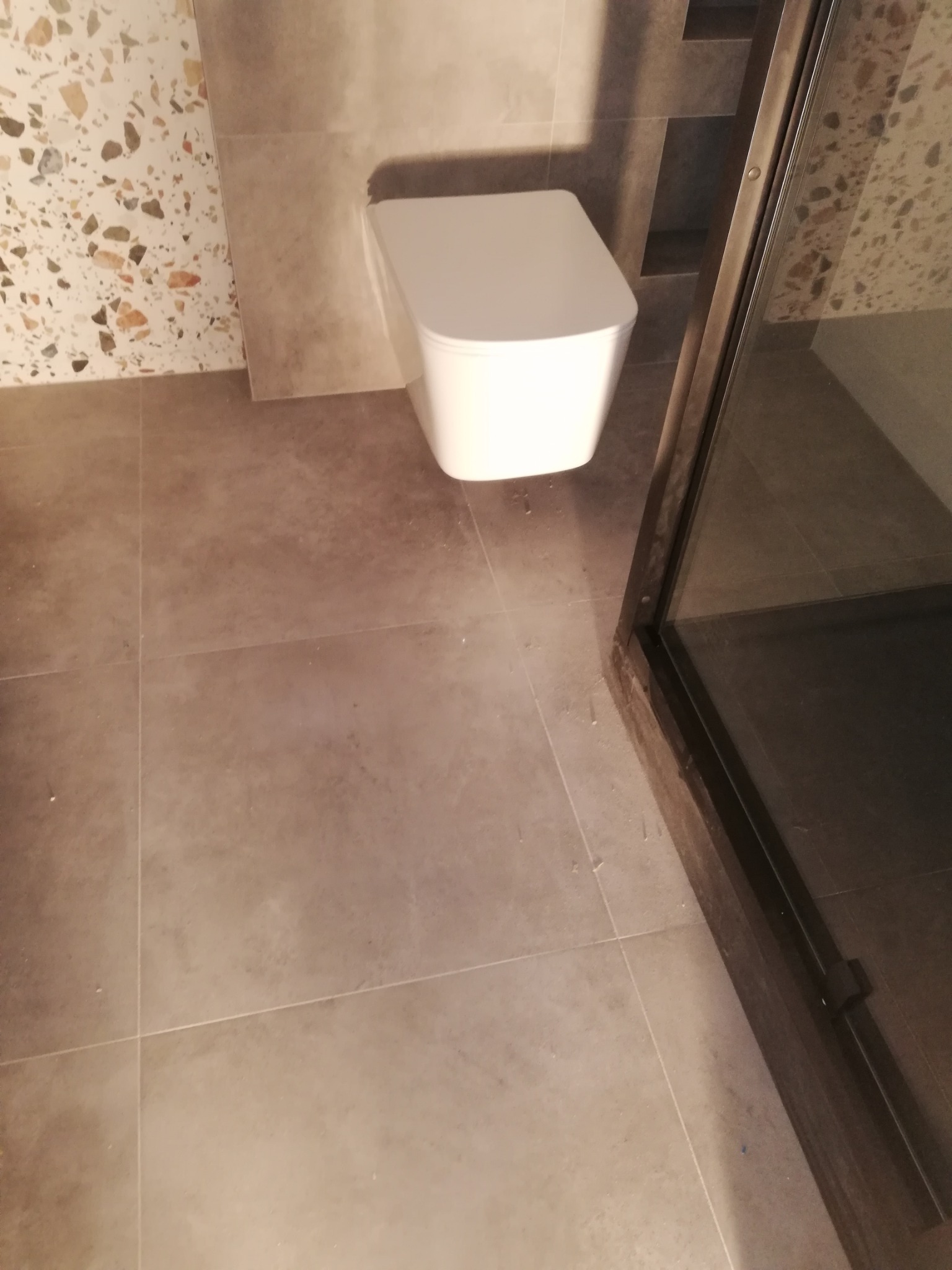 Nowoczesna toaleta podwieszana z deską wolnoopadającą, szare płytki podłogowe i ściany z mozaiką, kabina prysznicowa z czarną ramą