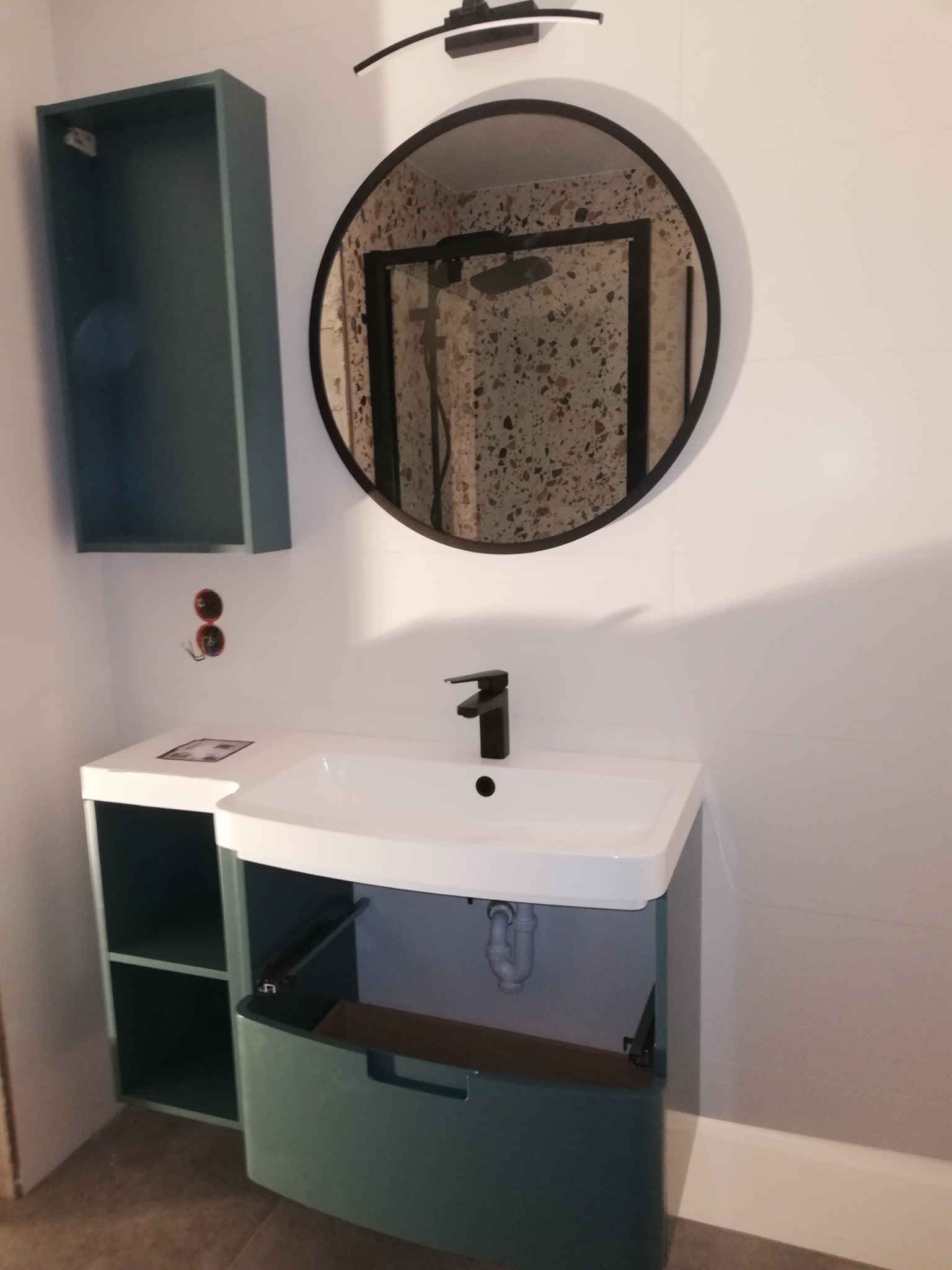 Nowoczesna łazienka z umywalką nablatową, okrągłym lustrem z czarną ramą odbijającym prysznic z mozaiką, wiszącą szafką i otwartą szafką pod umywalką w kolorze zielonym.