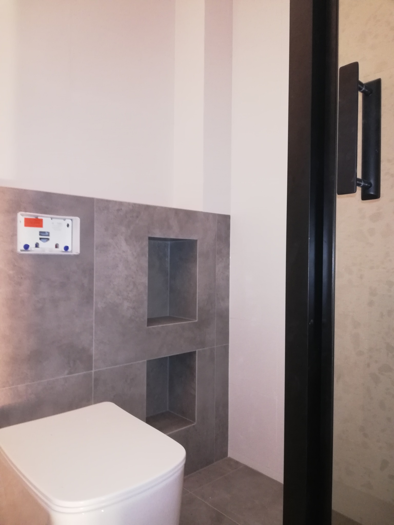 Nowoczesna łazienka z szarą, matową glazurą, wbudowanymi półkami w ścianie i wiszącą miską WC z widoczną instalacją podtynkową, oddzielona czarną ramą kabiny prysznicowej.
