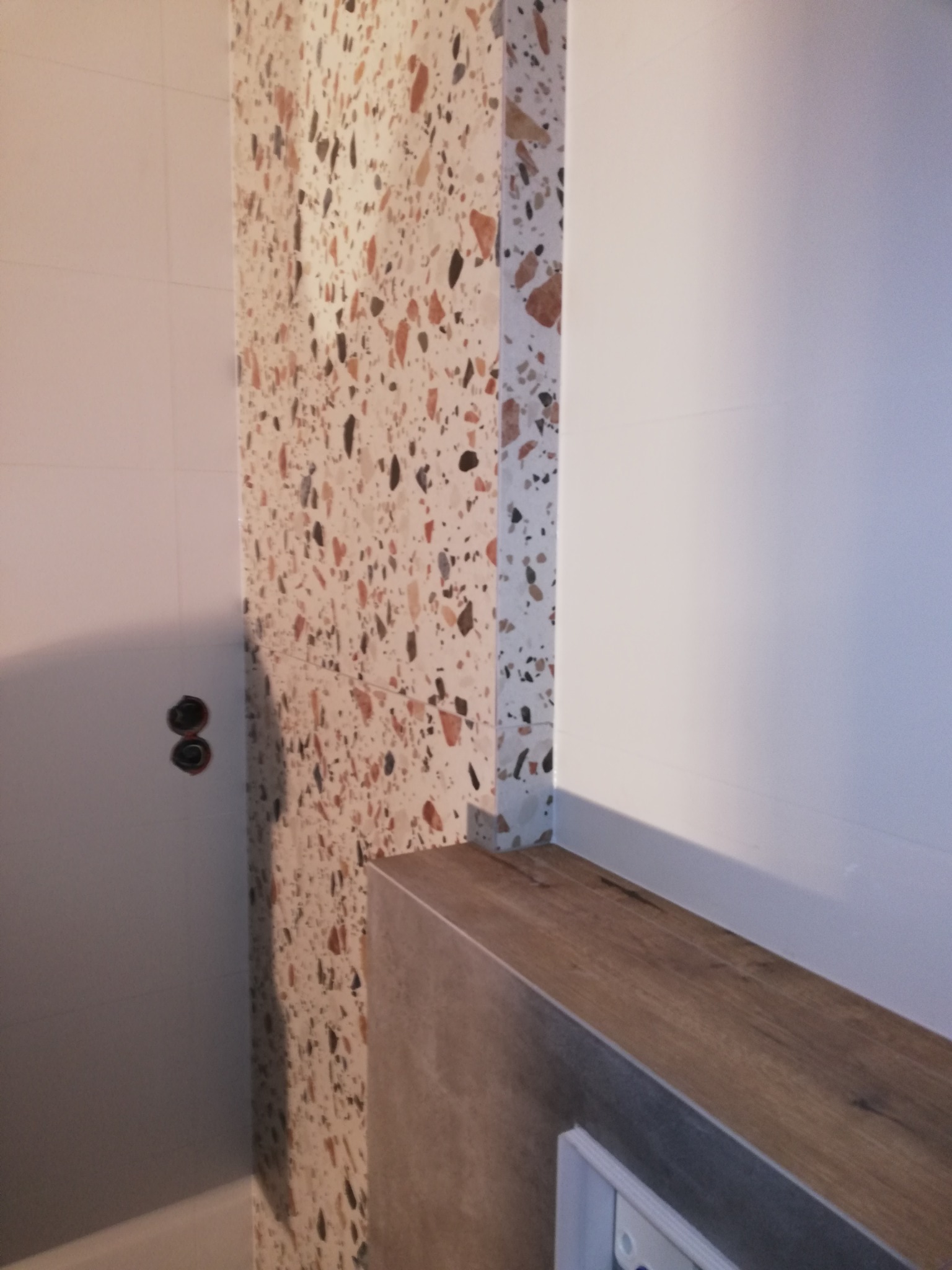 Fragment łazienki z wykończoną ścianą w płytkach lastryko, widoczne gniazdka elektryczne, zabudowa pod umywalkę z blatem imitującym drewno i białe płytki na ścianie.