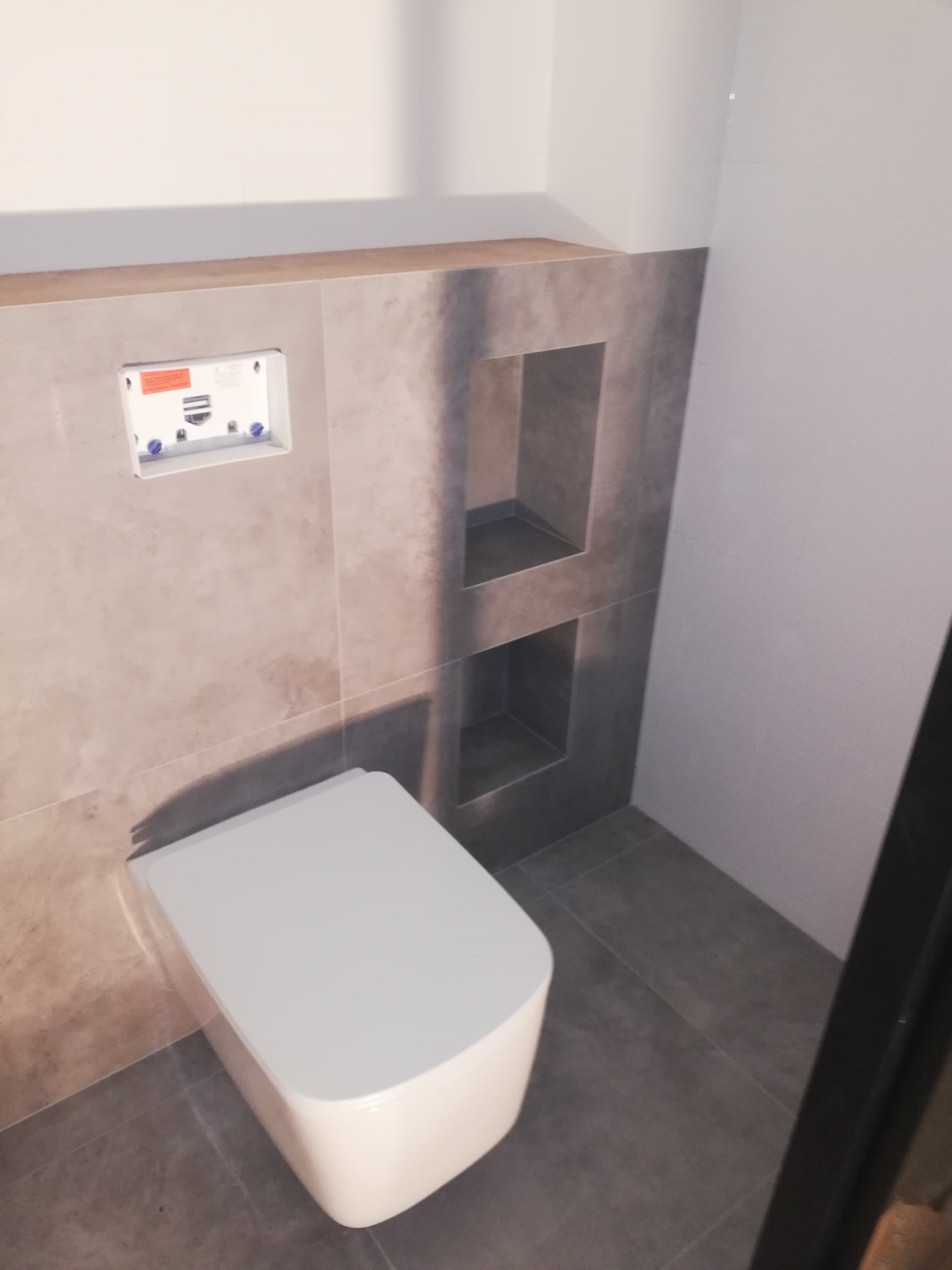 Nowoczesna toaleta podwieszana z szarą płytką imitującą kamień i wnękami w ścianie, widoczny stelaż podtynkowy.