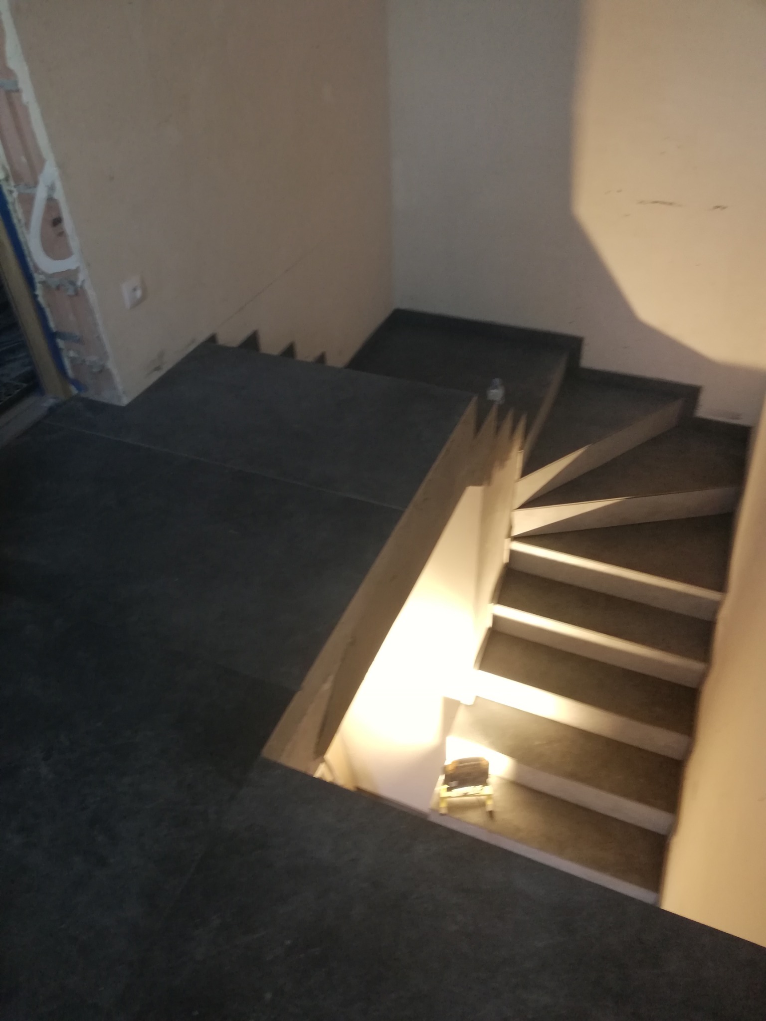 Schody wyłożone ciemnymi płytkami, widoczne ślady prac wykończeniowych na ścianach, oświetlenie robocze na schodach.