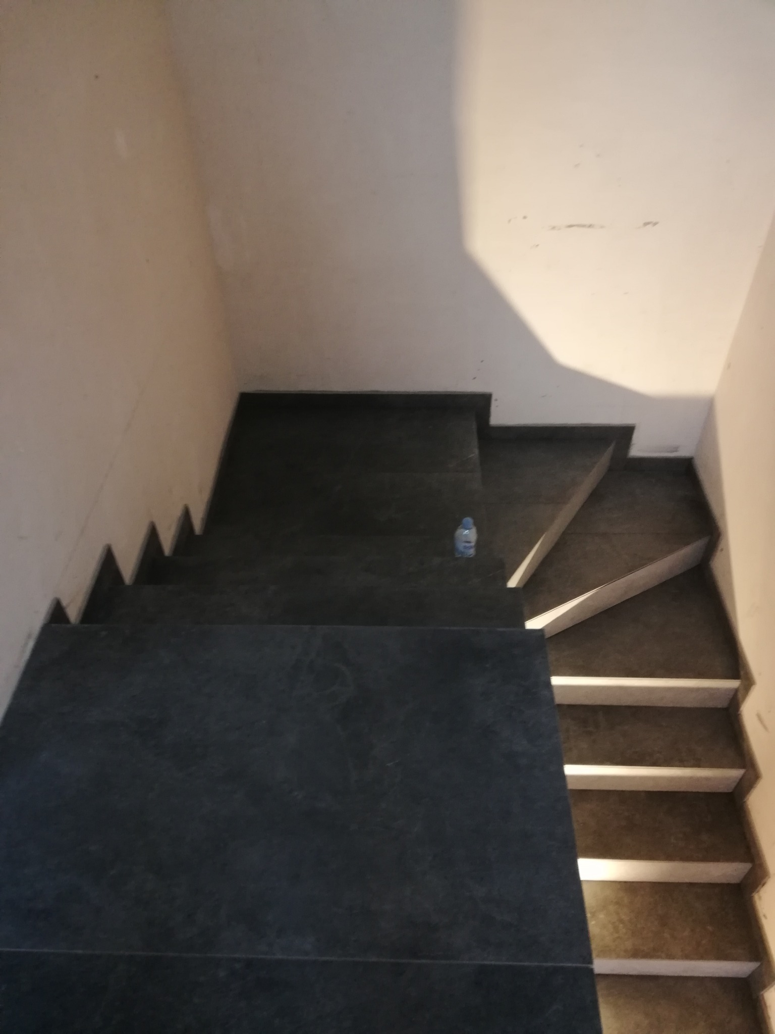 Schody wyłożone ciemną płytką, widok z góry, z butelką wody na jednym ze stopni. Część stopni wykończona białym pasem.