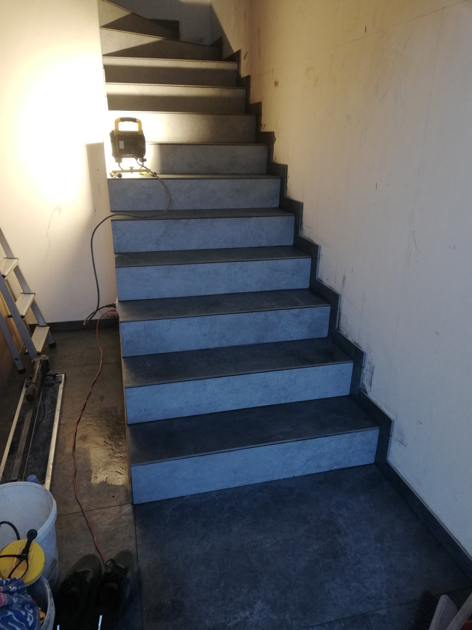 Schody wyłożone szarymi płytkami, oświetlone lampą budowlaną, z widocznymi narzędziami i częścią drabiny w tle, ukazujące etap prac wykończeniowych.