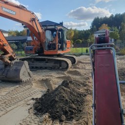 Przedsiebiorstwo sprzetowo transportowe Jarosław Sznajder - Pomarańczowa koparka Doosan na piaszczystym terenie, widoczne ślady gąsienic i mała pryzma ciemnej ziemi, w tle zabudowa jednorodzinna i błękitne niebo z chmurami.