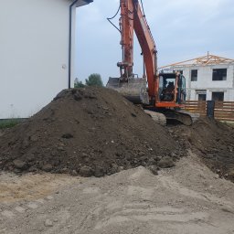 Przedsiebiorstwo sprzetowo transportowe Jarosław Sznajder - Pomarańczowa koparka Doosan stoi na hałdzie ziemi, w tle budynek w stanie surowym i fragment białego budynku z rynnami.