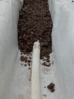 Biała rura drenażowa ułożona w wykopie wyłożonym białą geowłókniną, wypełnionym częściowo brązowym żwirem.