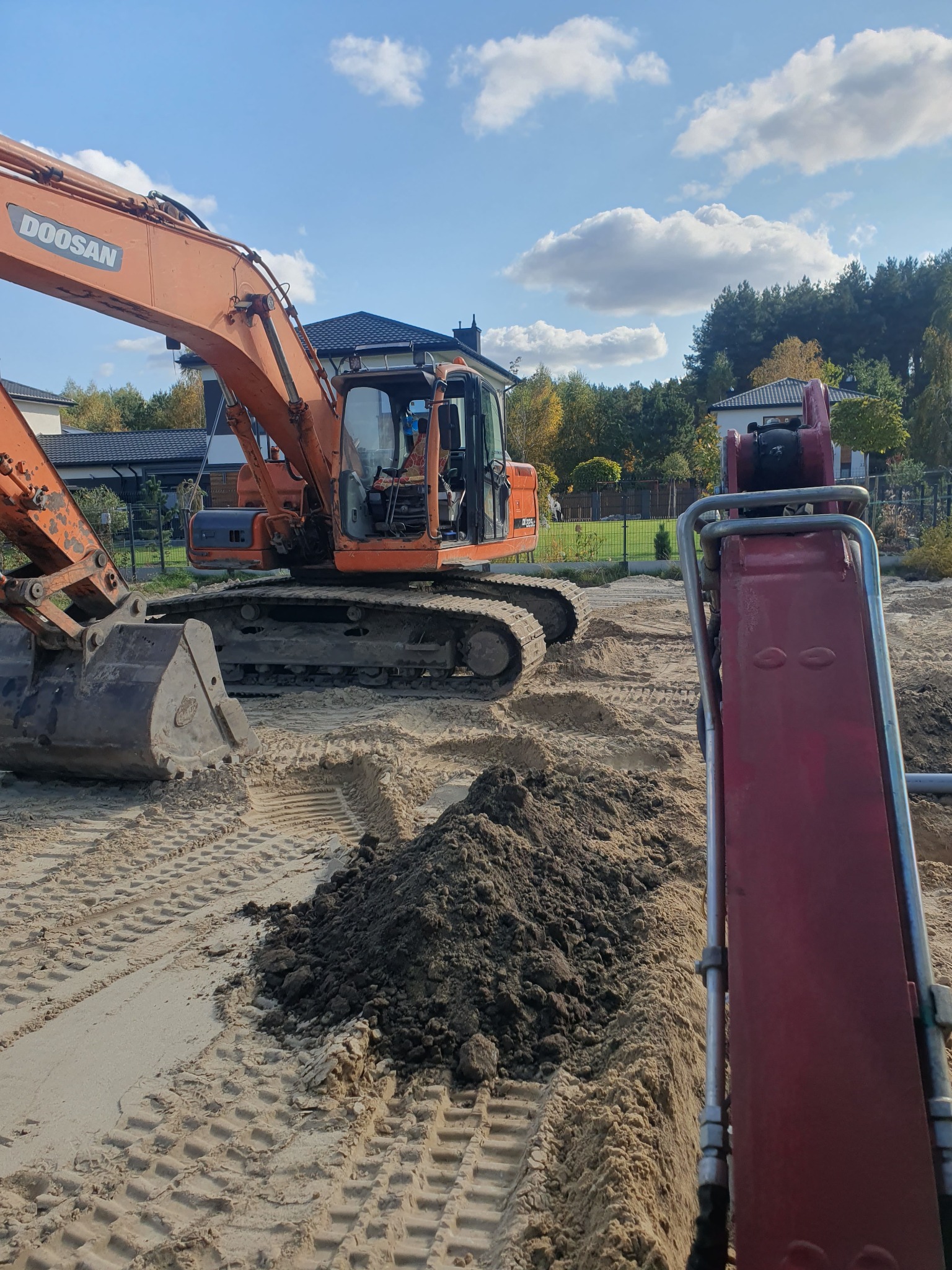 Pomarańczowa koparka Doosan na piaszczystym terenie, widoczne ślady gąsienic i mała pryzma ciemnej ziemi, w tle zabudowa jednorodzinna i błękitne niebo z chmurami.