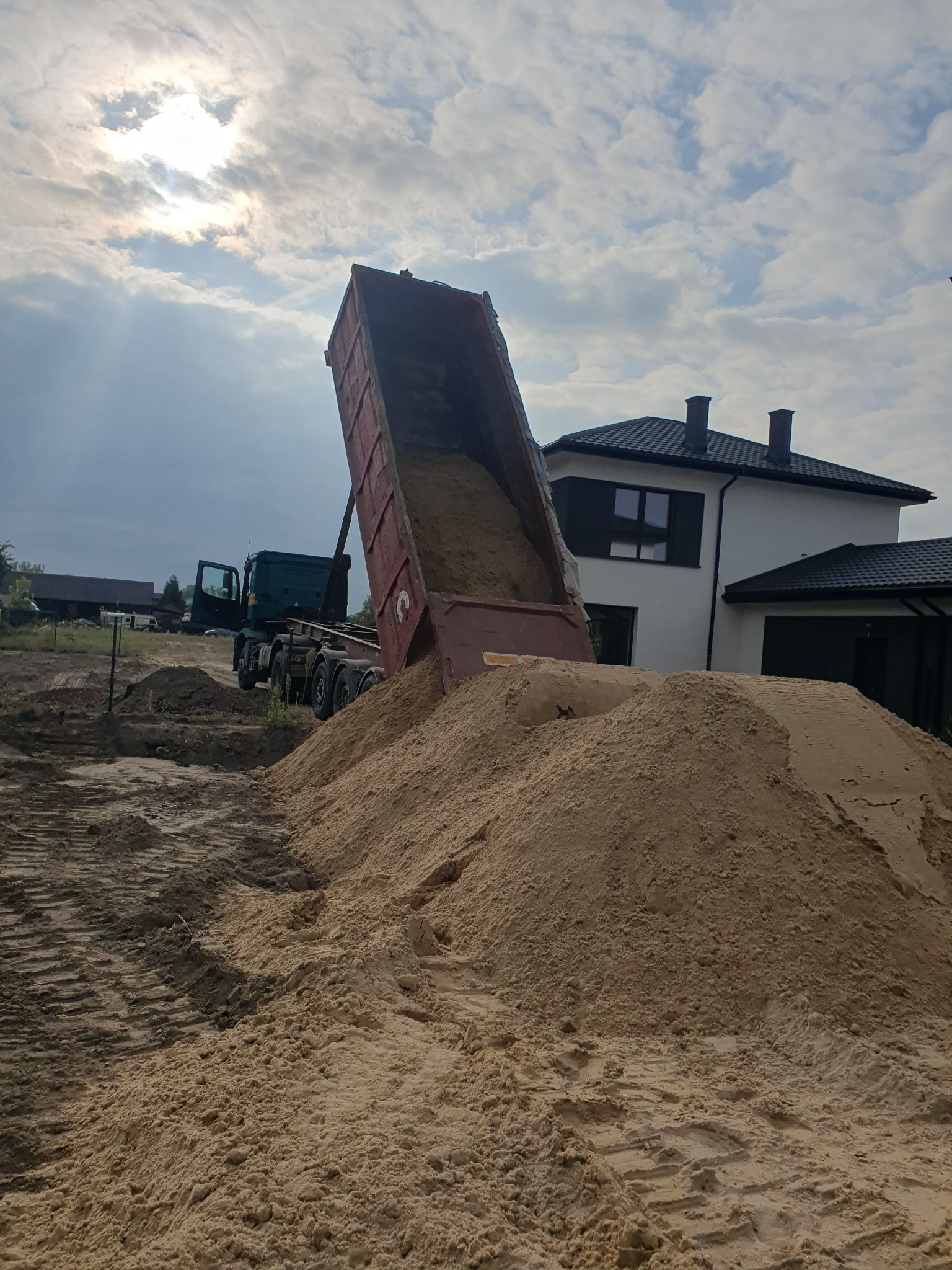 Wyładowywanie piasku z wywrotki na placu budowy przed nowoczesnym domem jednorodzinnym, widoczne ślady opon na ziemi.