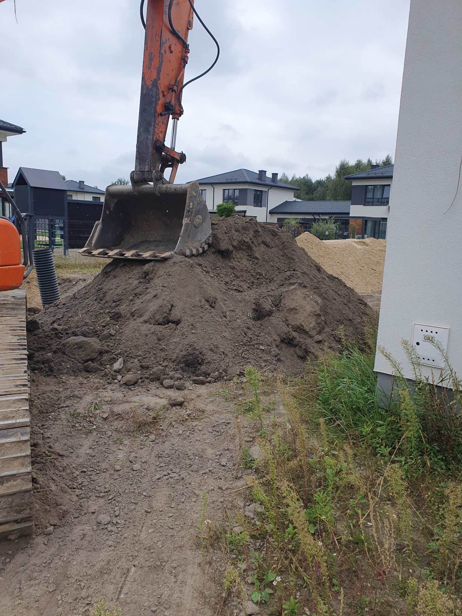 Pomarańczowa koparka z łyżką nad stertą ziemi na placu budowy, w tle domy jednorodzinne i stos piasku.