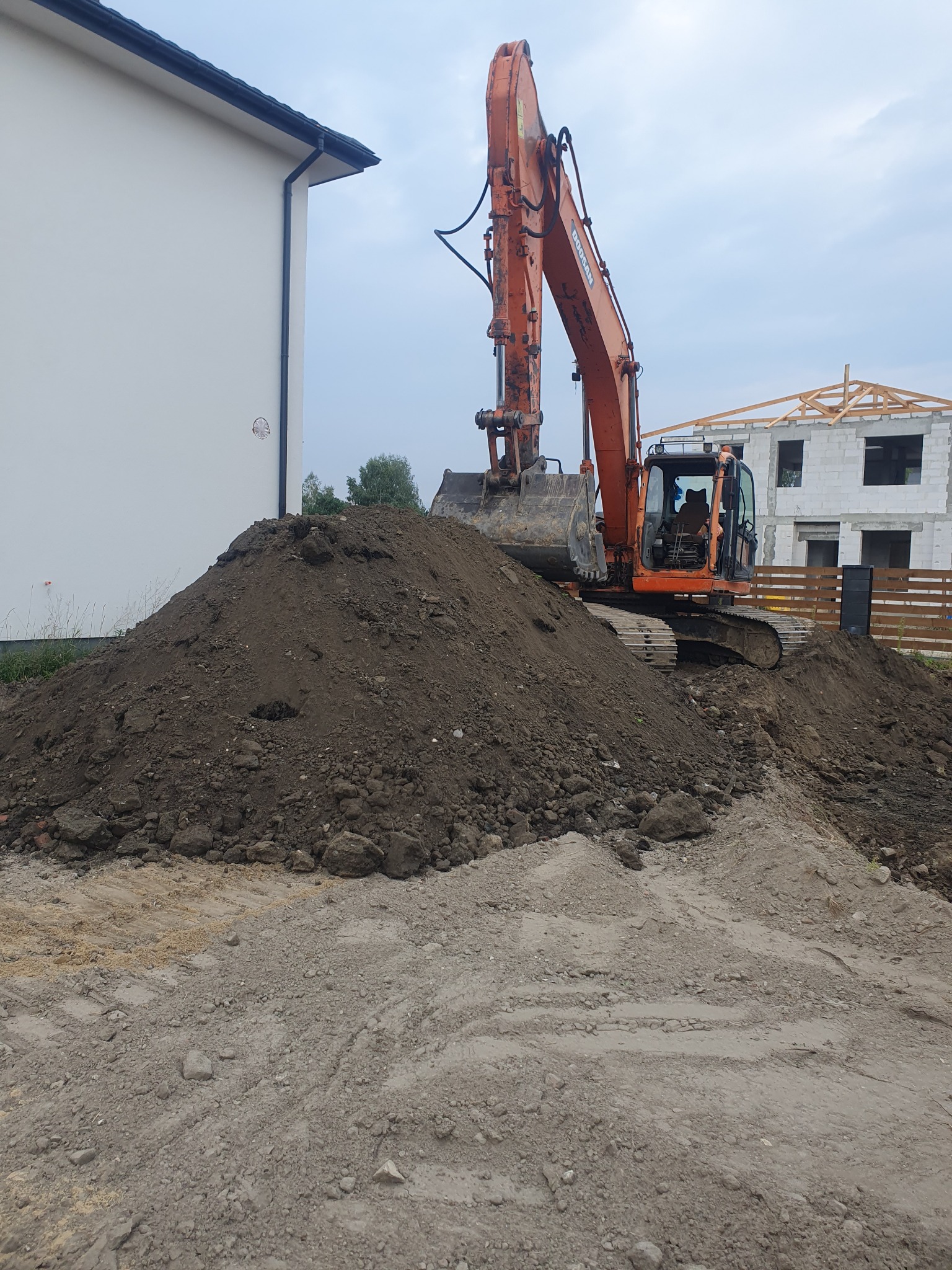Pomarańczowa koparka Doosan stoi na hałdzie ziemi, w tle budynek w stanie surowym i fragment białego budynku z rynnami.
