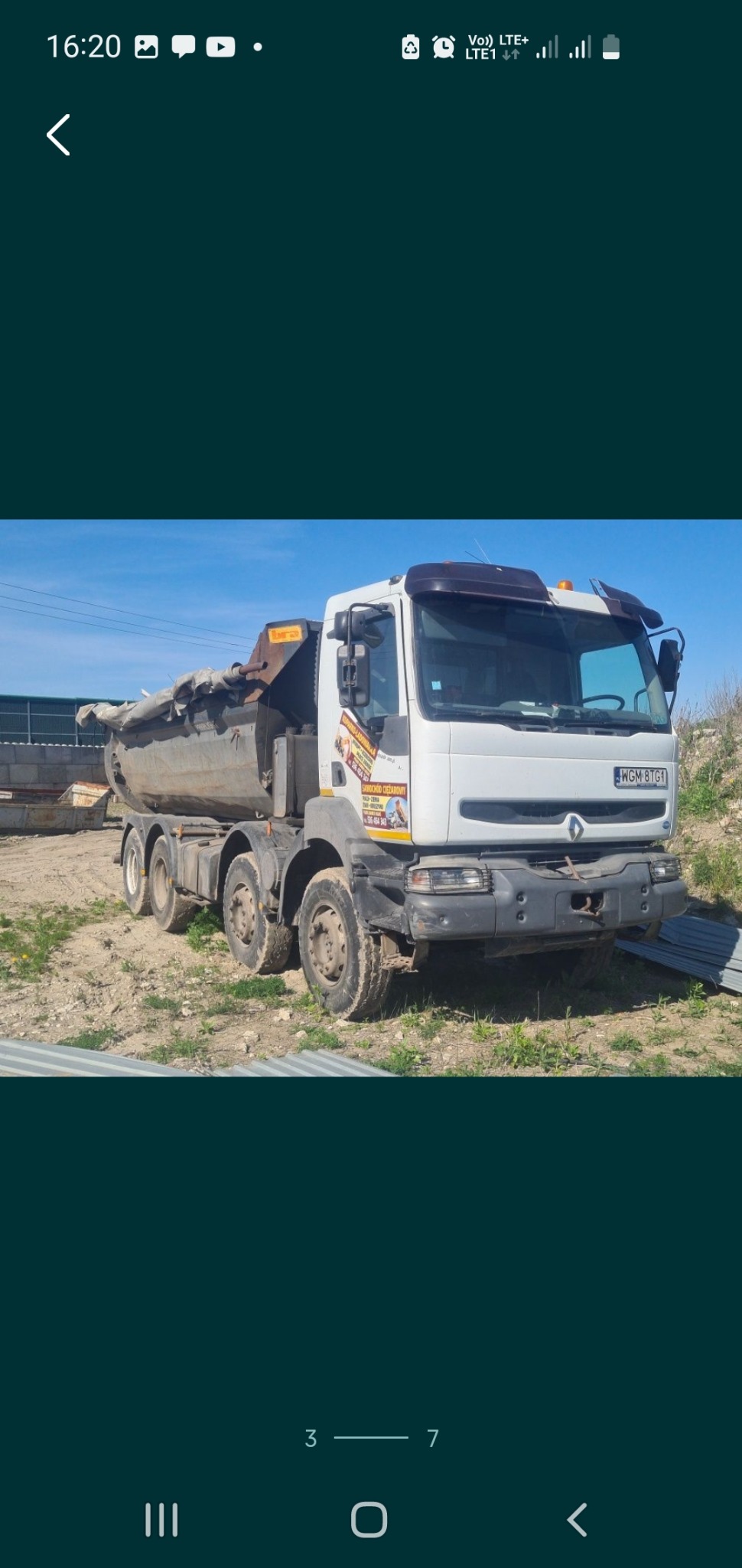 Ciężarówka wywrotka Renault z częściowo zakrytą plandeką na terenie budowy, w tle ogrodzenie z blachy falistej i błękitne niebo.