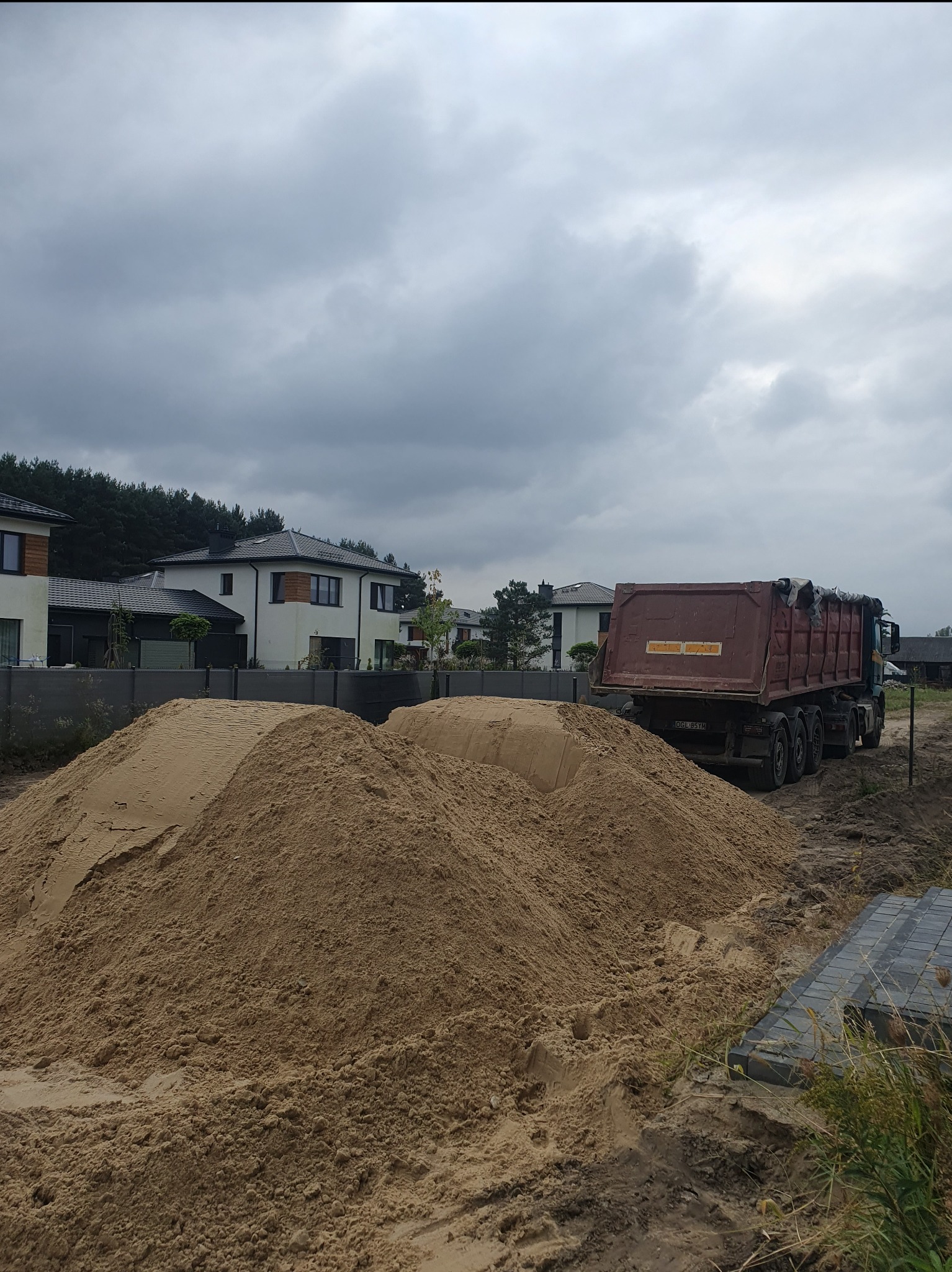 Ciężarówka wywrotka z otwartą skrzynią stoi obok dużej pryzmy piasku na placu budowy, w tle nowoczesne domy jednorodzinne i pochmurne niebo.