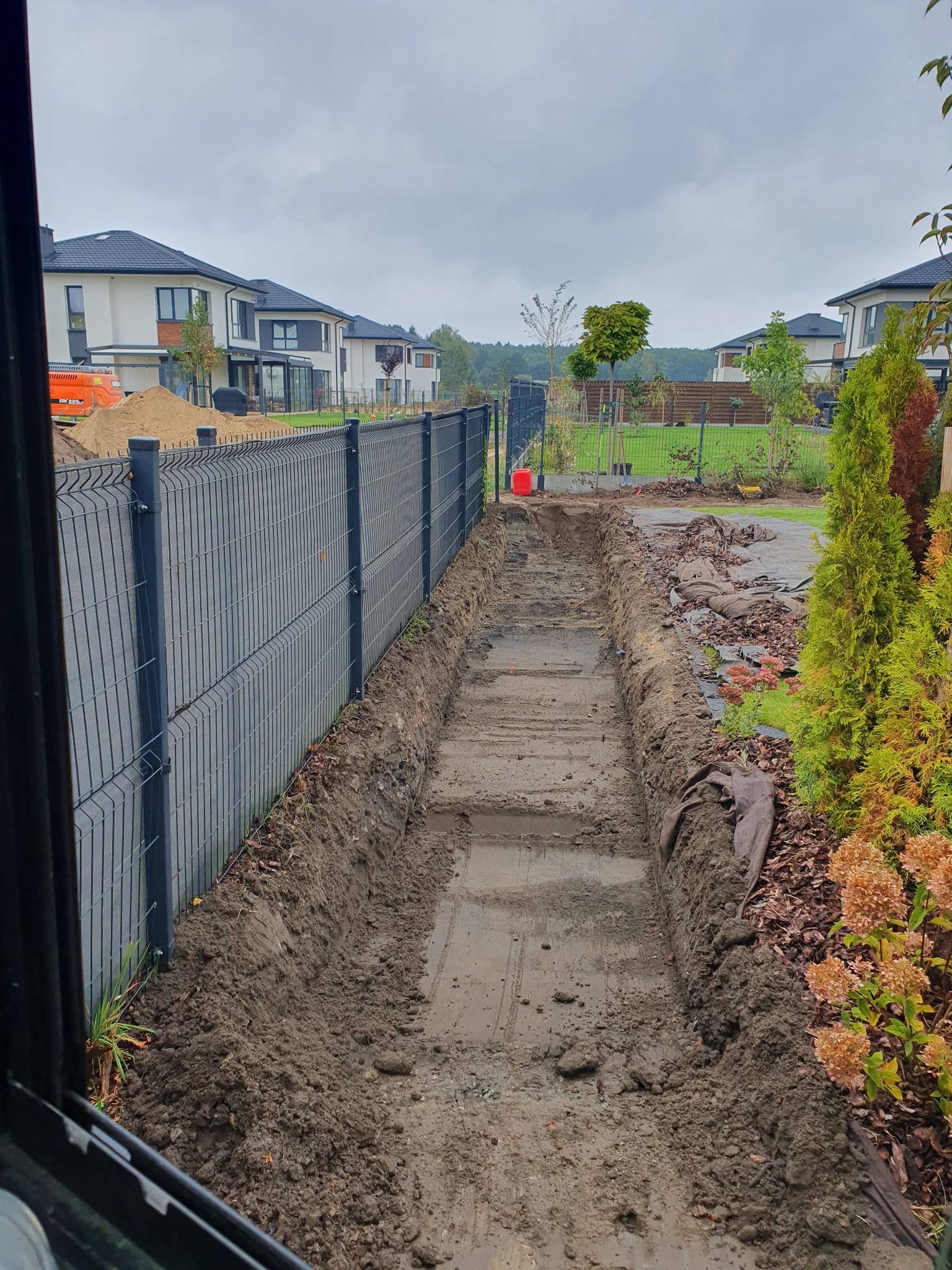 Wykopany wąski rów wzdłuż metalowego ogrodzenia panelowego na posesji z domami jednorodzinnymi w tle, widok z wnętrza kabiny maszyny.