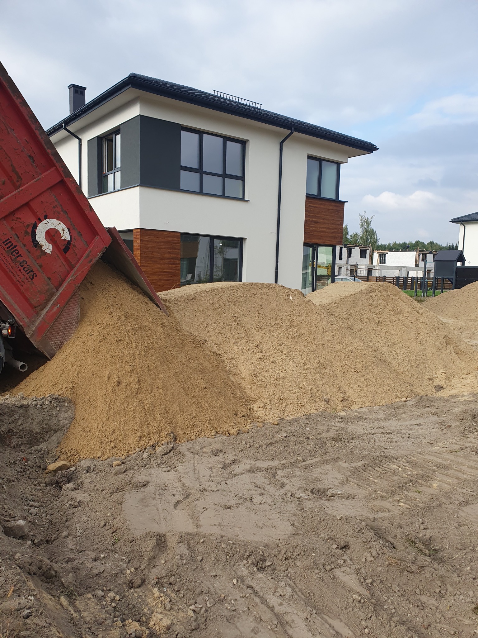 Czerwona wywrotka z logiem Inter Cars wysypuje piasek na plac budowy, w tle nowoczesny dom jednorodzinny z szarymi elementami i budynek w trakcie wznoszenia z białego pustaka.
