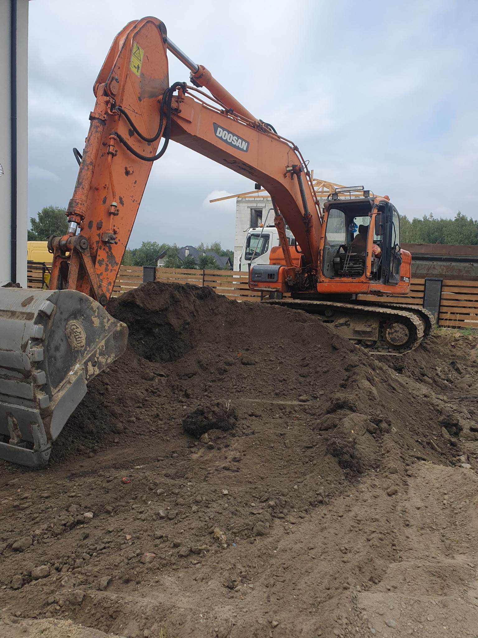 Pomarańczowa koparka Doosan nabiera ziemię na tle budowy domu jednorodzinnego z widoczną konstrukcją dachu.