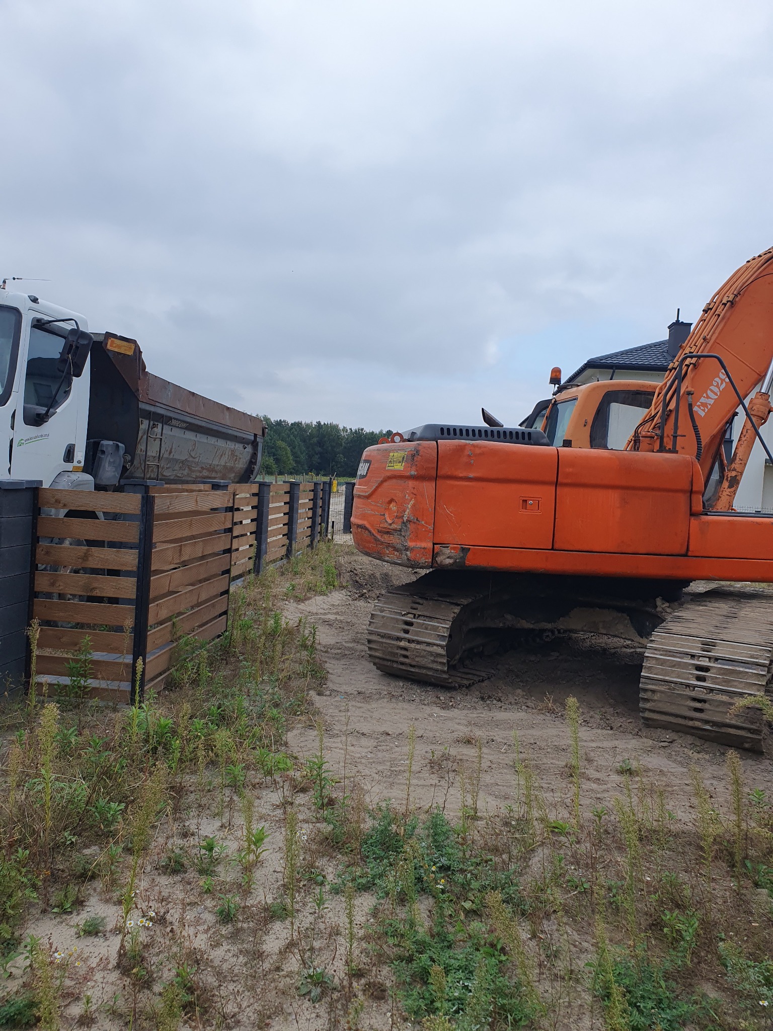 Pomarańczowa koparka gąsienicowa i biała wywrotka obok drewnianego ogrodzenia na piaszczystym terenie budowy, widok z boku.