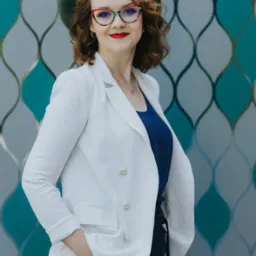 Uśmiechnięta kobieta z rudymi włosami w okularach w ozdobnych oprawkach, ubrana w białą marynarkę i granatową bluzkę, pozuje na tle dekoracyjnej ściany w odcieniach błękitu i szarości.