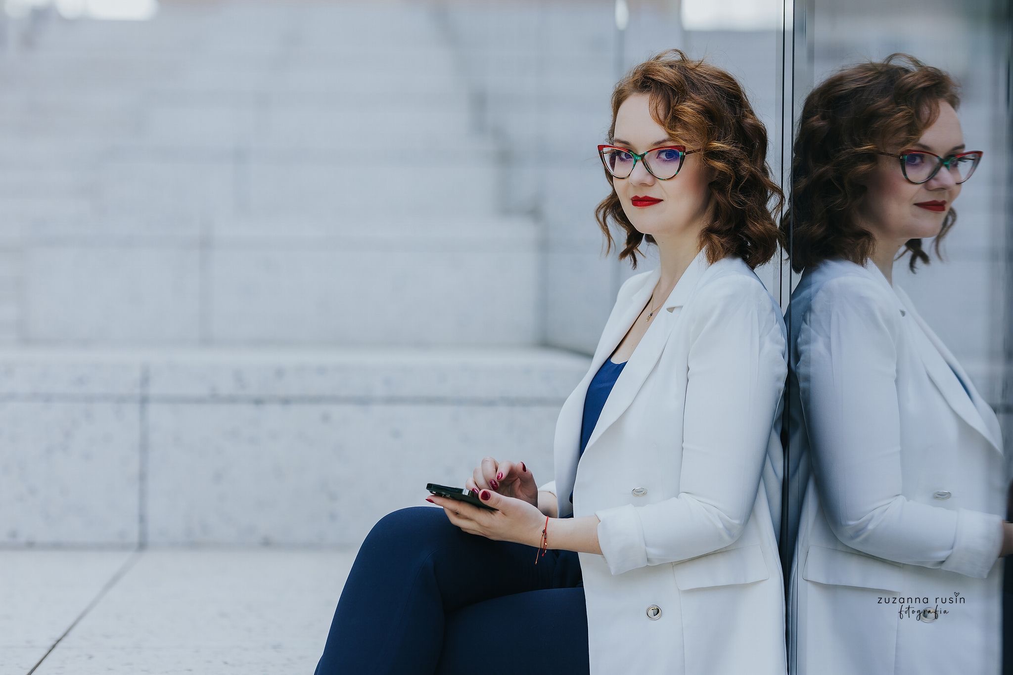 Portret kobiety w okularach i białym żakiecie, siedzącej na tle schodów i szklanej ściany z odbiciem.